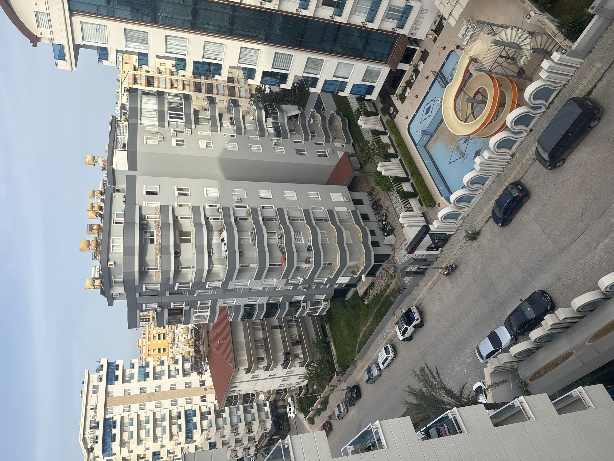 Here is a detailed description of the image:
The image shows an aerial view of a densely populated urban area, likely a resort city. The scene is dominated by multiple high-rise buildings of varying architectural styles and heights. The buildings predominantly feature balconies and windows, indicating residential or hotel purposes.
In the middle of the frame is a swimming pool area with a water slide. Along a side street are several parked cars.
The sky in the background suggests a clear or partly cloudy day. The image captures the scene in daylight.