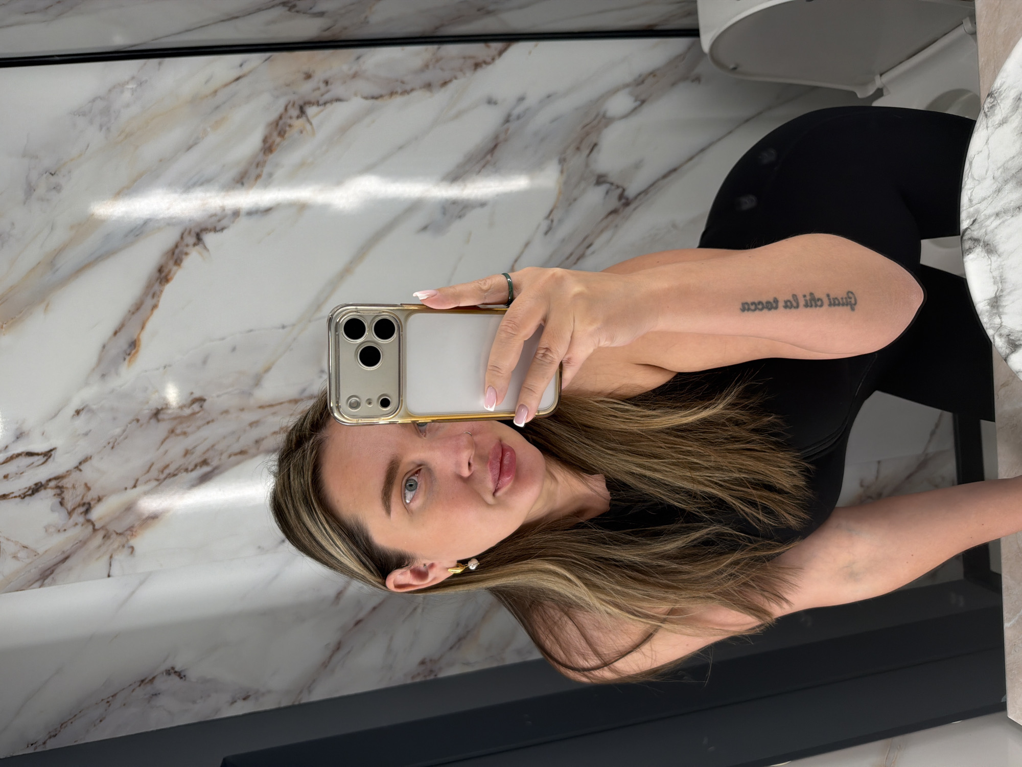 A woman with shoulder-length highlighted hair is taking a selfie in a bathroom. She is wearing a black top and black leggings. Her phone is a modern iPhone with a clear case. She is wearing a ring on her left hand. A tattoo is visible on her arm. The bathroom features marble-patterned walls and countertop and black trim. A toilet is partially visible in the background. The photo does not contain sufficient information to identify the location in Moscow.