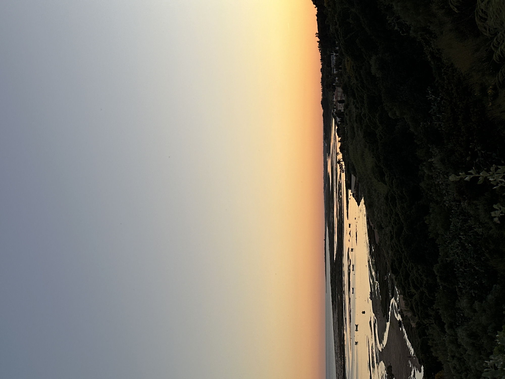 The image depicts a twilight or sunset scene, possibly at the mouth of the Guadiana River bordering Vila Real de Santo António, Portugal. The sky transitions from a light blue-grey to a warm orange. The river or estuary is visible on the right, with patches of exposed sand and boats dotting the water's surface. Lush, dark greenery covers the riverbank, interspersed with a few buildings.