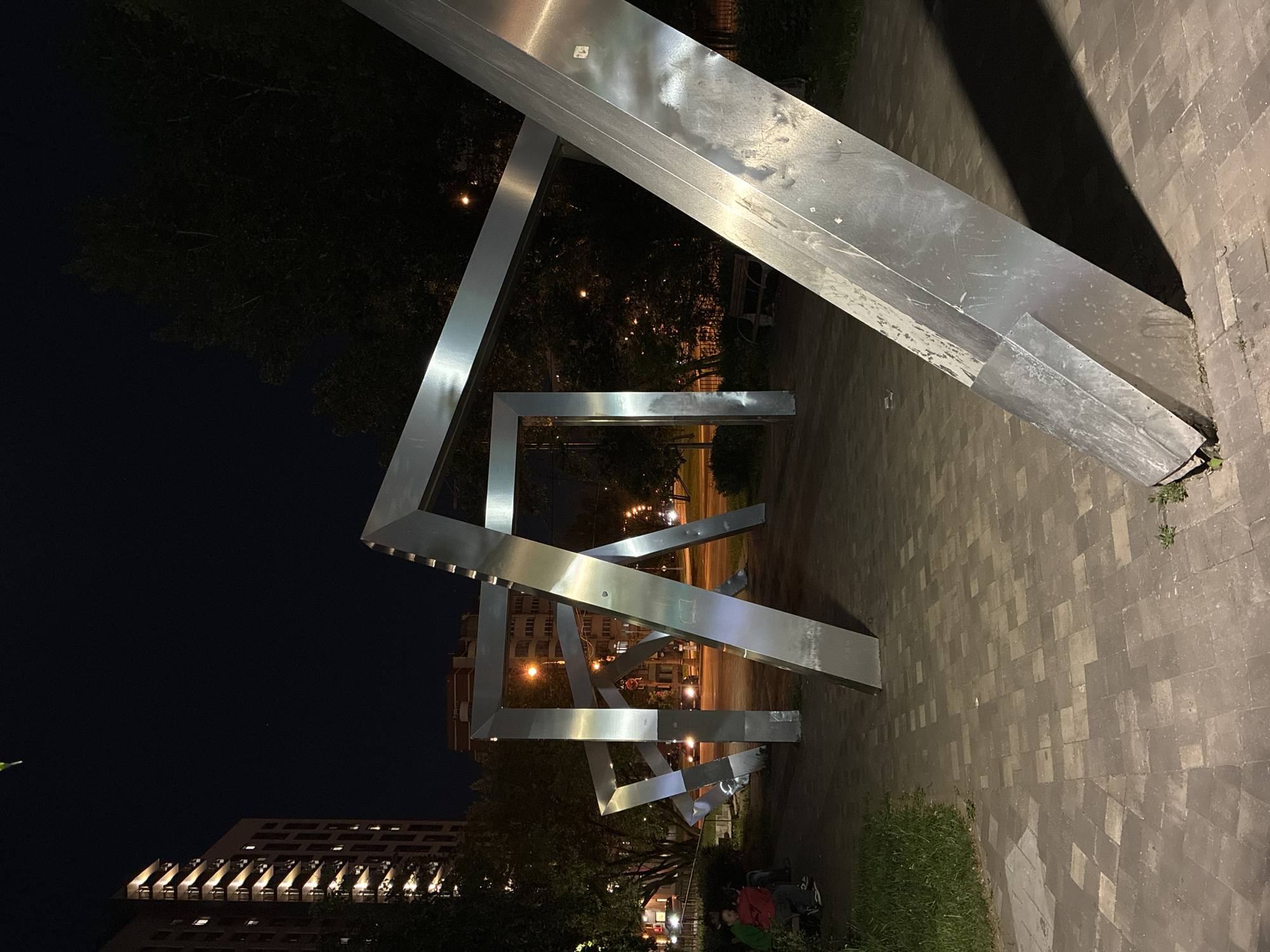 At night, a public art installation in the city of Moscow, Russia, is illuminated. The sculpture consists of a series of intersecting, angular, silver-colored metal beams. A paved walkway leads to the installation, with greenery visible on either side. In the background, urban architecture and streetlights are seen. A group of people is positioned near the base of the sculpture.