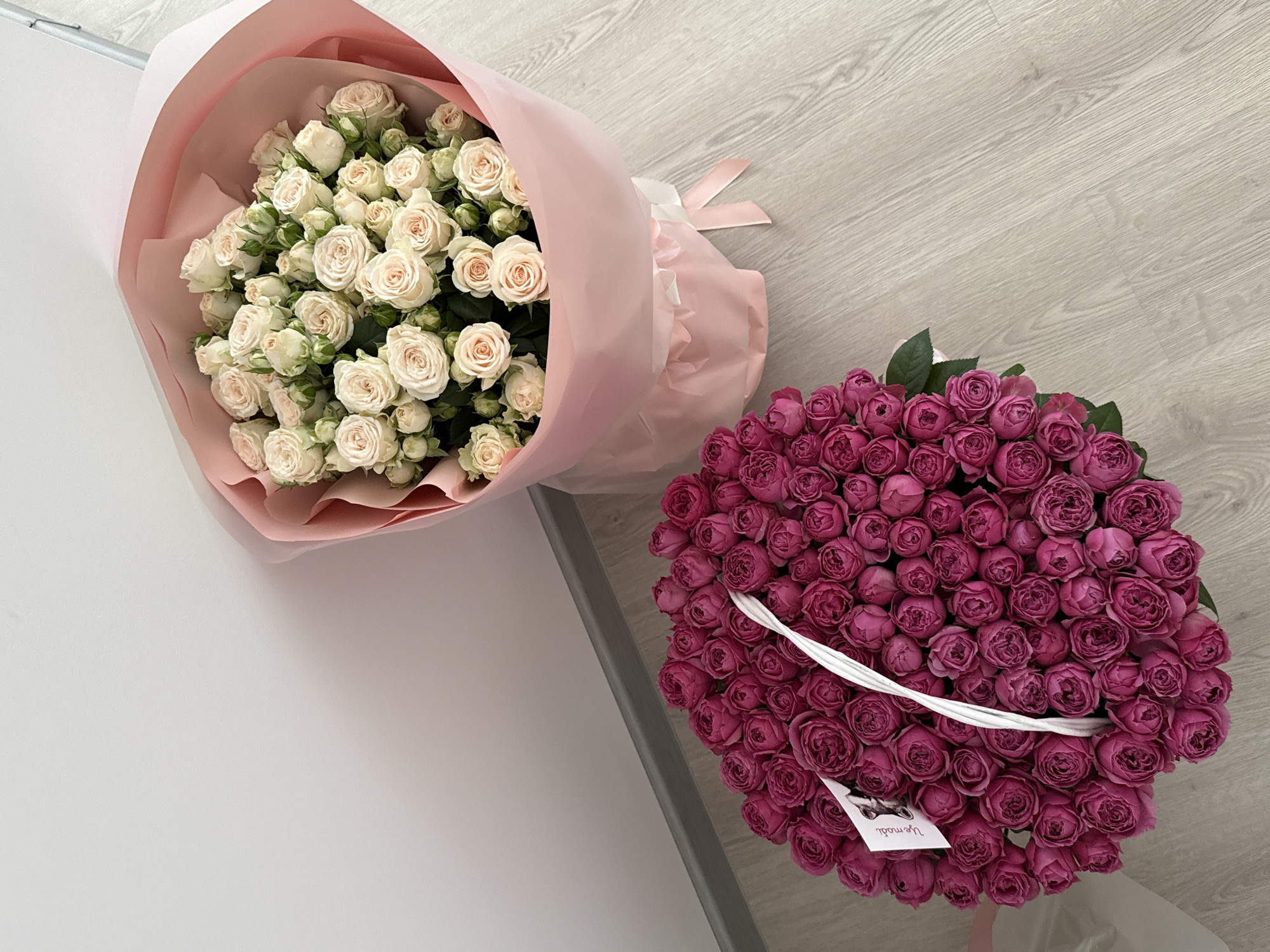 Two bouquets of roses are displayed on a light-colored wooden floor against a white wall. The bouquet on the left is larger and contains numerous deep pink roses packed tightly together in a white basket. A small white card with a dog illustration and text is attached to the handle of the basket. The bouquet on the right is smaller and features a collection of light cream-colored roses wrapped in pink paper. The scene is devoid of people and shows no indication of a specific urban location.