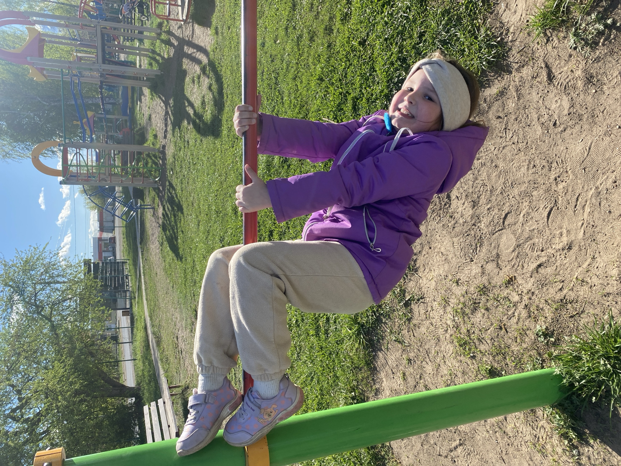 A young girl is hanging on a horizontal bar at a playground. She wears a purple jacket, light-colored pants, and purple shoes. She also has a light-colored headband. The playground has various play structures made of metal and plastic. The ground is covered with dirt and grass. The location is likely a playground in the city of Chaikovsky, Russia. The photo was taken in daylight with a clear sky.