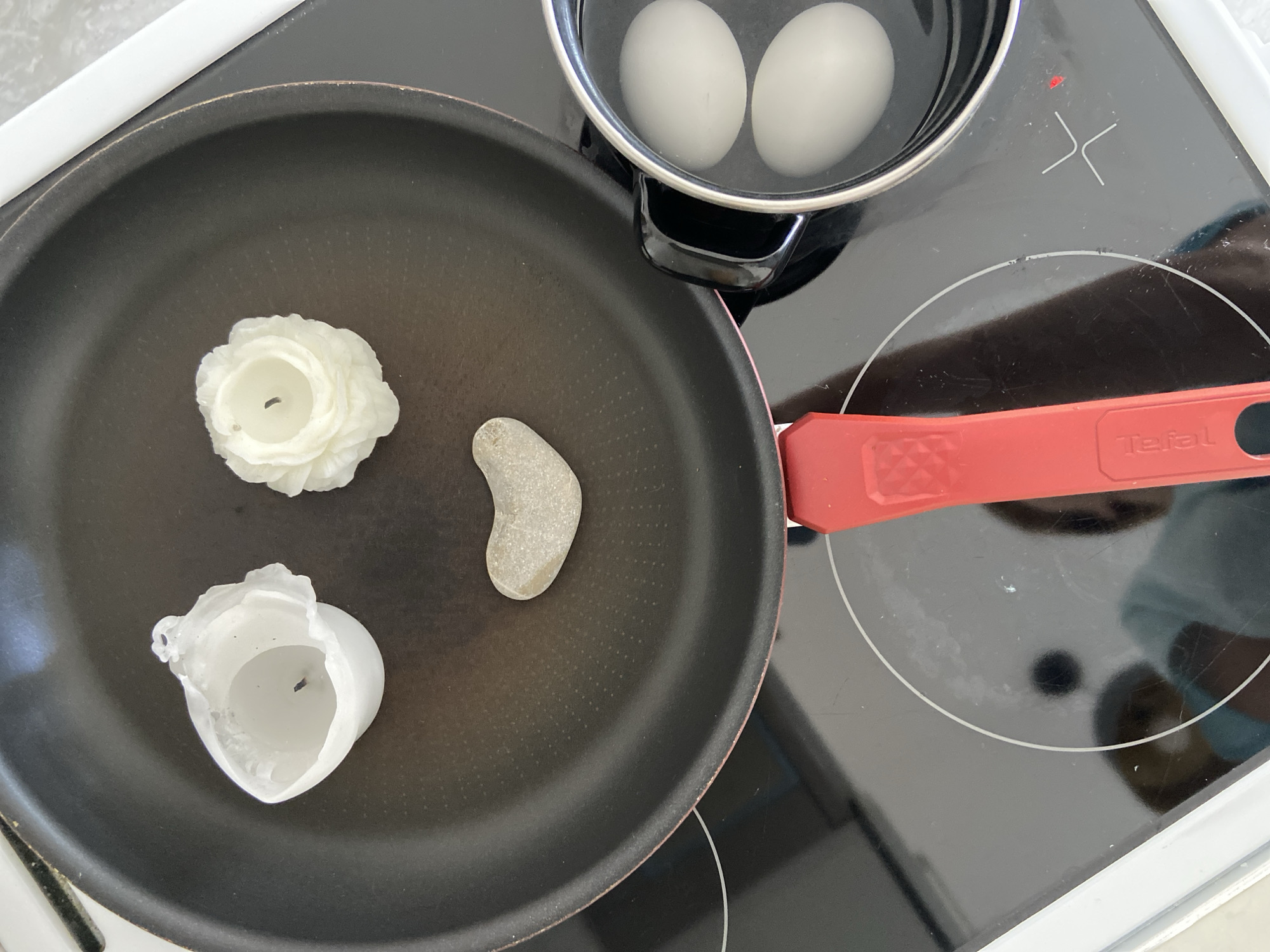 The scene is a close-up, overhead shot of a stovetop with a frying pan and a saucepan. The frying pan contains two white candle holders and a smooth, heart-shaped stone. The saucepan, placed on a lit burner, contains two white eggs submerged in water. The frying pan has a red handle with the word "Tefal" printed on it. The stovetop is black and reflects the surrounding environment, including a person wearing a face mask. The location is not evident from the image, making it impossible to confirm if it is in Budva, Montenegro.