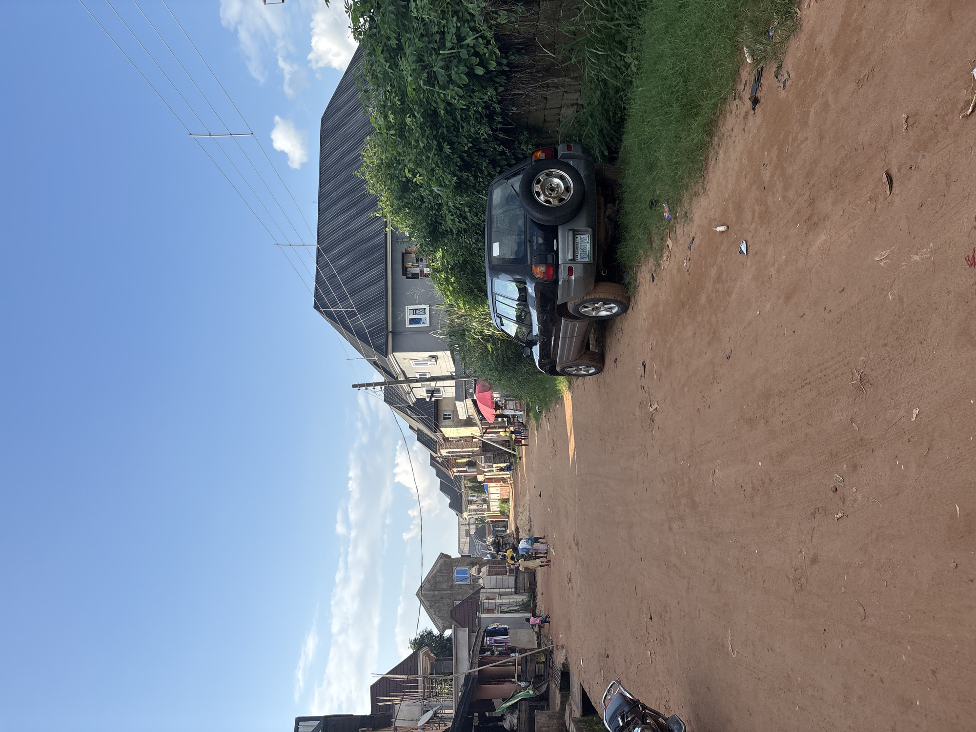 In Obosi, Nigeria, a dirt road stretches through a residential area lined with multi-story buildings. A black SUV is parked on the right side of the road, partially obscured by lush green foliage. Several individuals, including children, are visible walking along the road and gathered in small groups. The sky above is a clear blue with a few scattered clouds. The scene suggests a typical day in a Nigerian neighborhood.