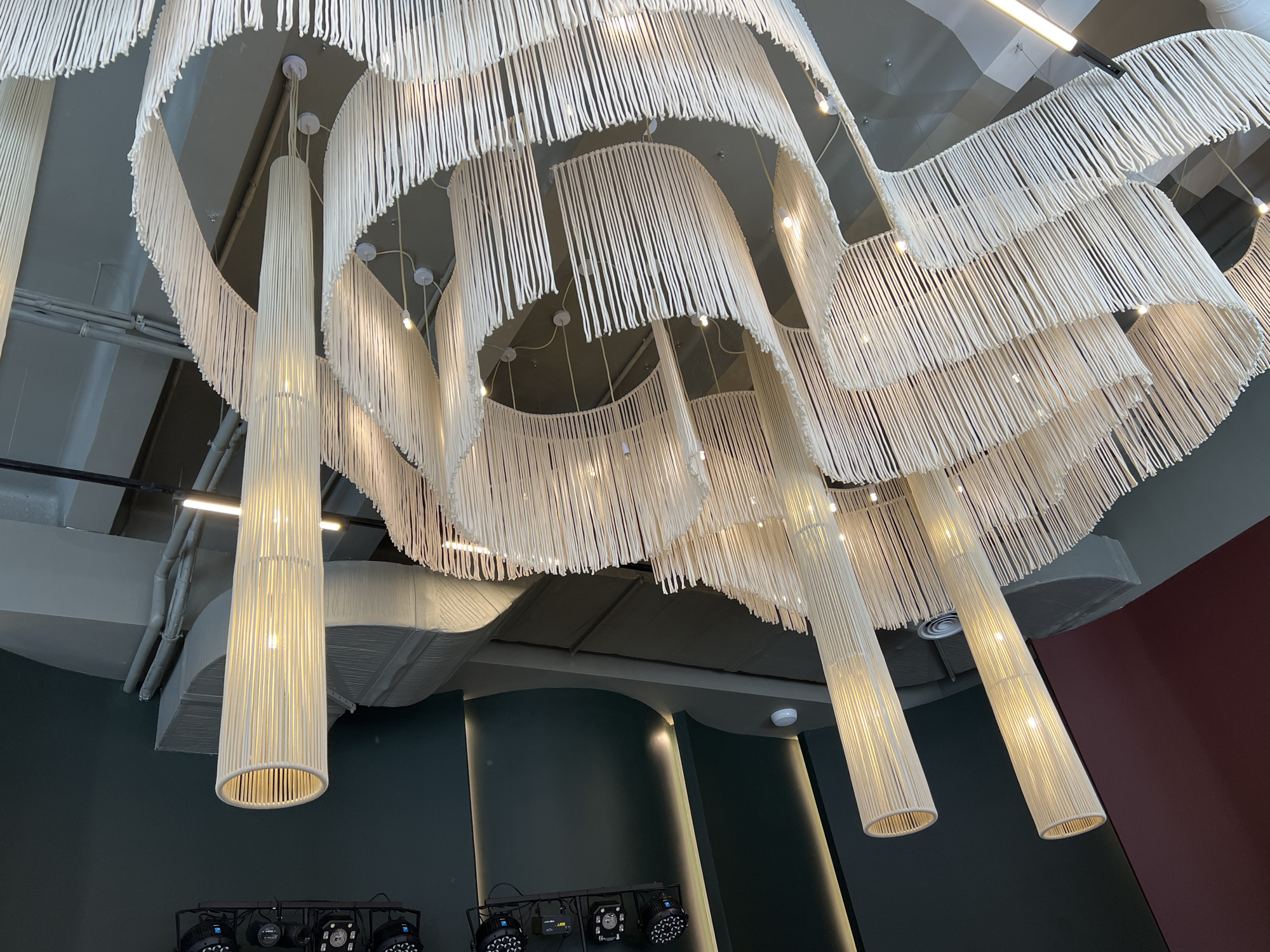 This is an indoor shot of a ceiling featuring a complex, layered lighting installation. The primary visual element consists of multiple tiers of white, fringe-like material forming large, flowing shapes suspended from the ceiling. Interspersed among these tiers are several elongated, cylindrical light fixtures made of the same white material, emitting a warm, ambient glow.
The setting appears to be a restaurant or event space with a modern design. The walls are painted in a combination of dark green and burgundy. Stage lighting fixtures are mounted on the lower section of the wall. Overhead, the ceiling is mostly gray and appears to incorporate industrial architectural elements such as ductwork and structural beams.
Based on the architectural style and decor, this location is potentially in Saint Petersburg, Russia.