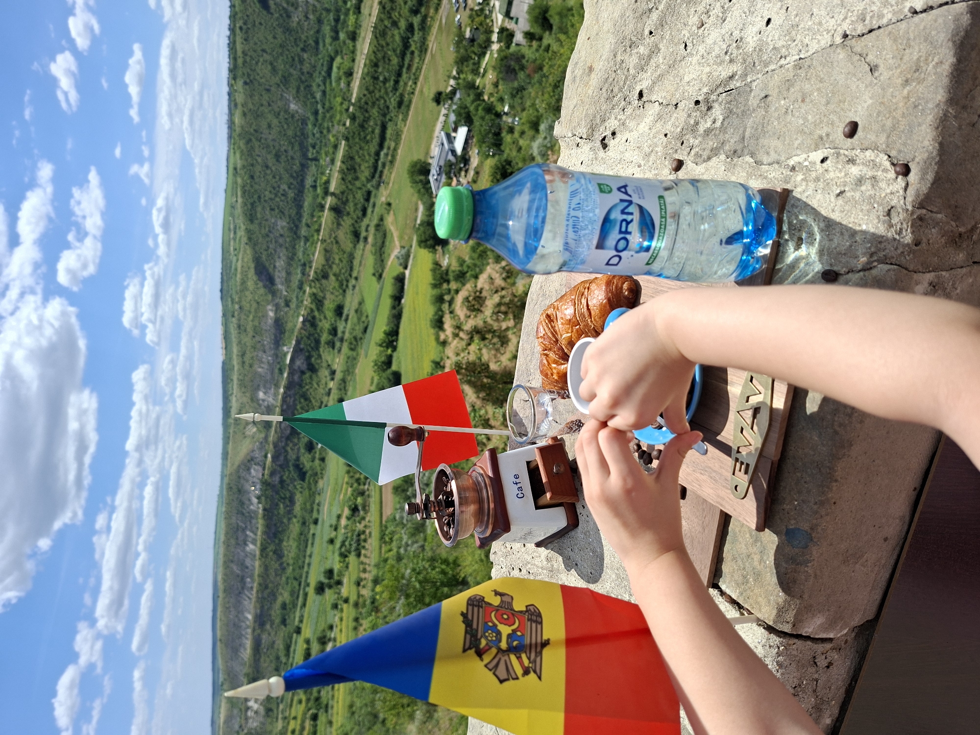 In Trebujeni, Moldova, on a stone ledge overlooking a verdant valley under a partly cloudy sky, a scene is set for a rustic breakfast. Two small flags, one the national flag of Moldova and the other the flag of Italy, stand upright. In front of them, a vintage-style manual coffee grinder sits next to a clear glass of water and a croissant. A bottle of "DORNA" mineral water with a green cap is positioned to the right. Below these items, a child's hands are actively arranging coffee beans on a small wooden board inscribed with the word "CEVAV". The background reveals a vast landscape with rolling green hills, a winding dirt road, and scattered buildings. The lighting suggests a bright, sunny day.