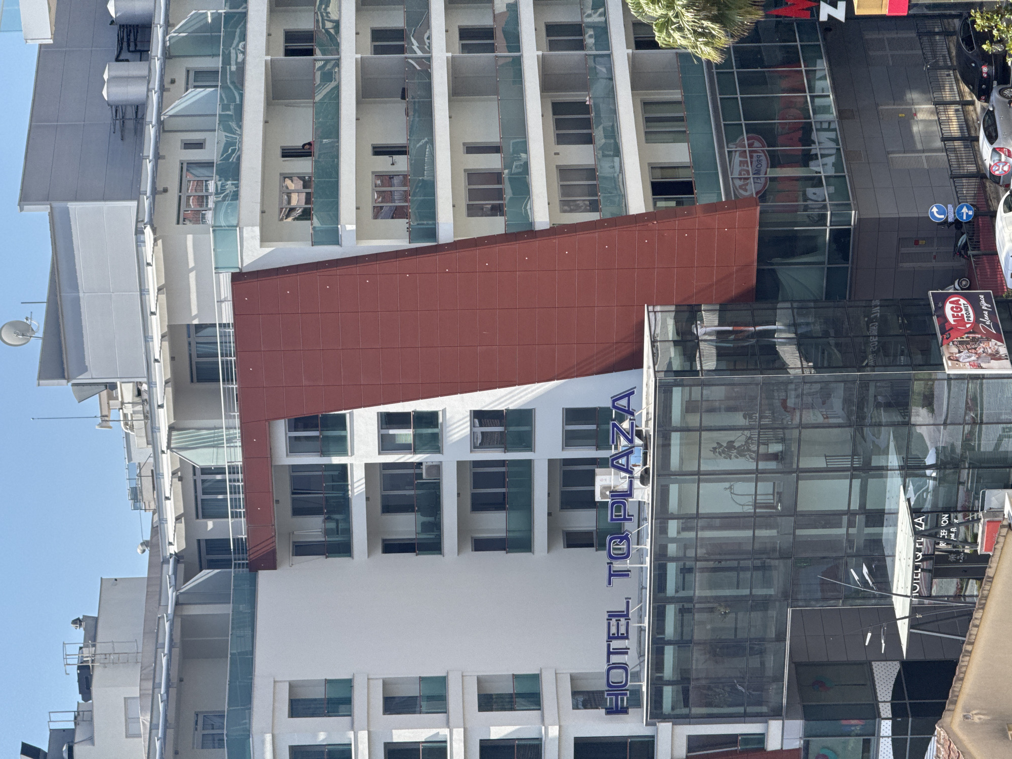 The image captures a modern, multi-story hotel named "HOTEL TO PLAZA" in Budva, Montenegro. The hotel features a prominent red, angled facade, contrasting with the white and glass exterior of the rest of the building. Several floors of the hotel have balconies with glass railings, some of which have people visible on them. A large glass-fronted section at the base of the hotel displays reflections of the surrounding environment and internal signage. The hotel is situated in a dense urban area, with other buildings visible in the background and foreground. Cars are parked along the street in front of the hotel, and a palm tree is visible on the right side of the image. The sky is clear and blue, indicating a sunny day.