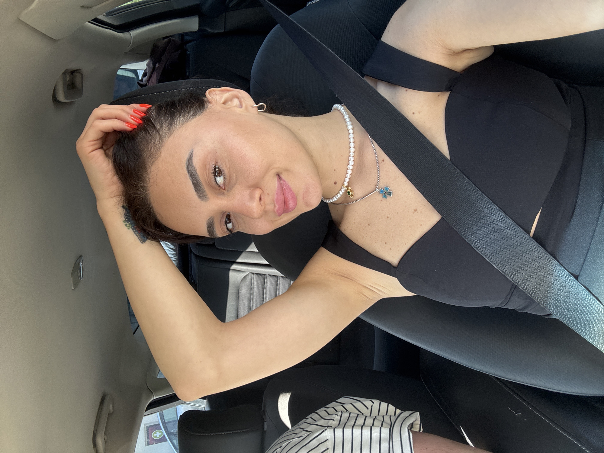 A young woman with dark hair, brown eyes, and light skin is captured in a selfie taken from inside a car. She is wearing a black sleeveless top and a pearl necklace with two pendants, one of which is a blue cross. Her right arm is raised, with her hand resting on the back of her head. Her fingernails are painted bright red. The woman is wearing a seatbelt. The car interior is visible, including the passenger seat and the back seats. The environment outside the car suggests it is daytime. The image appears to be taken in Tbilisi, Georgia, based on the visible street signs with Georgian writing.