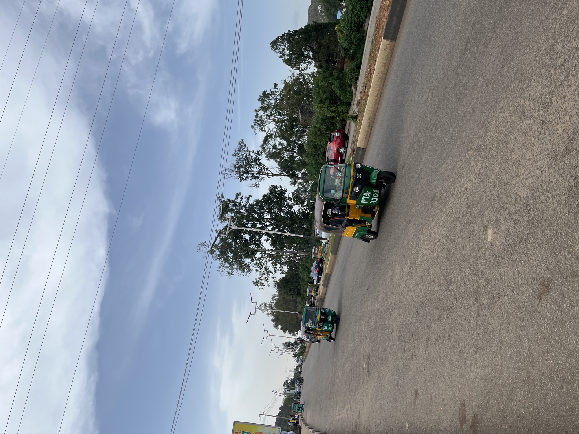 This image depicts a street scene in Gura Top, Nigeria, likely during the daytime. The perspective is from a low angle, looking upwards at the sky and then down the road.
The main subjects are several auto-rickshaws, also known as "keke napeps," driving on a paved road. These are colorful, three-wheeled vehicles, predominantly green and yellow. One keke prominently displays "PTA 8301" in white lettering. Some of the kekes appear to have passengers inside, suggesting it's a mode of public transportation.
In the background, there are lush green trees and vegetation lining the side of the road, indicating a somewhat natural environment interspersed with urban infrastructure. A red car is visible parked or stopped on the side. Overhead, a network of power lines stretches across the sky, which is a mix of blue with white clouds. The sky suggests it's daytime, and the weather appears clear.
The overall impression is of a typical street scene in a Nigerian town, with active transportation and everyday life unfolding. There are no strong emotional cues, but the activity of the kekes suggests movement and commerce. The visible text on the keke is the license plate number.