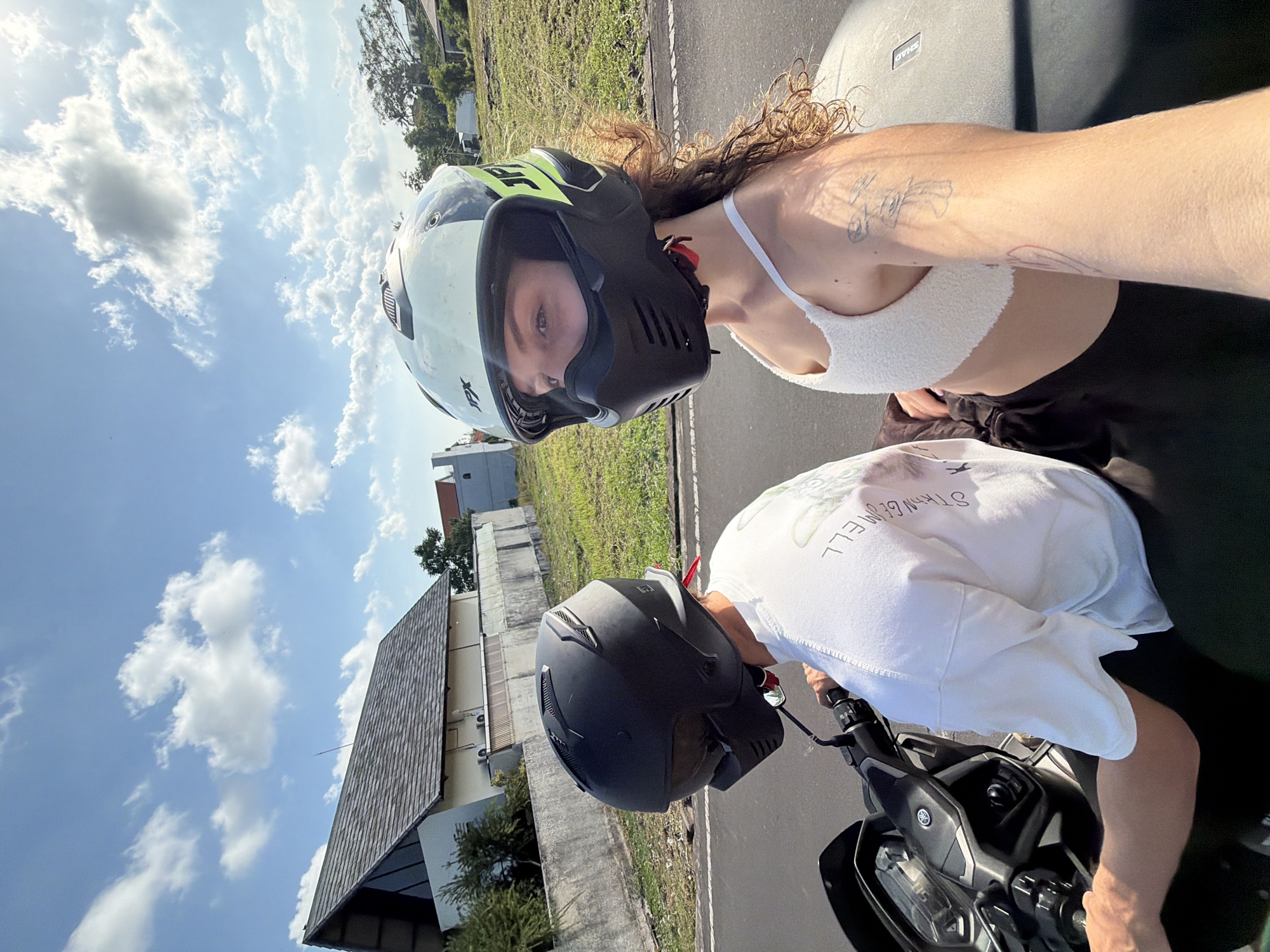 In a medium, eye-level shot, two individuals are seated on a scooter on a paved road with a sidewalk and grassy embankment to the right. The person in the foreground, on the right, is facing the camera and wearing a white cropped top and black bottoms. They have curly brown hair and are wearing a light-colored motorcycle helmet with green accents. A tattoo of three mushroom-like figures is visible on their right arm. The person behind them, on the left, is facing forward and wearing a white t-shirt with text on the back and a black motorcycle helmet. The scooter is dark gray with a Yamaha logo on the front. The background shows a residential area with houses, some with peaked roofs, and a clear blue sky with scattered white clouds. The location is identified as North Kuta, Indonesia.