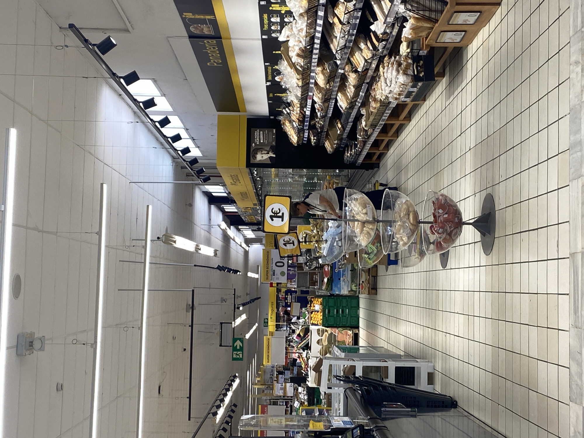 The image shows the interior of a supermarket in Bel Air, Spain. The perspective is from an aisle looking towards the front of the store, which is brightly lit by fluorescent and track lighting. The floor is tiled with large, square, off-white tiles. On the left, there are shelves with what appear to be frozen goods or storage units. Further down the aisle on the left, there are displays of fruits and vegetables, including oranges in green crates and watermelons. Signs for "SUPER ECONOMICO" are visible.
To the right, a large display of baked goods, identified by a "Panaderia" sign, stretches along the wall. The shelves are filled with various types of bread and pastries packaged in plastic bags. A woman in a uniform, likely an employee, is visible behind a counter in the middle of the store, interacting with products. In the foreground on the right, there are clear glass bowls displaying what look like small jars of preserves or condiments. Yellow signage with prices like "1€" is interspersed throughout the displays. The overall environment suggests a well-stocked grocery store.