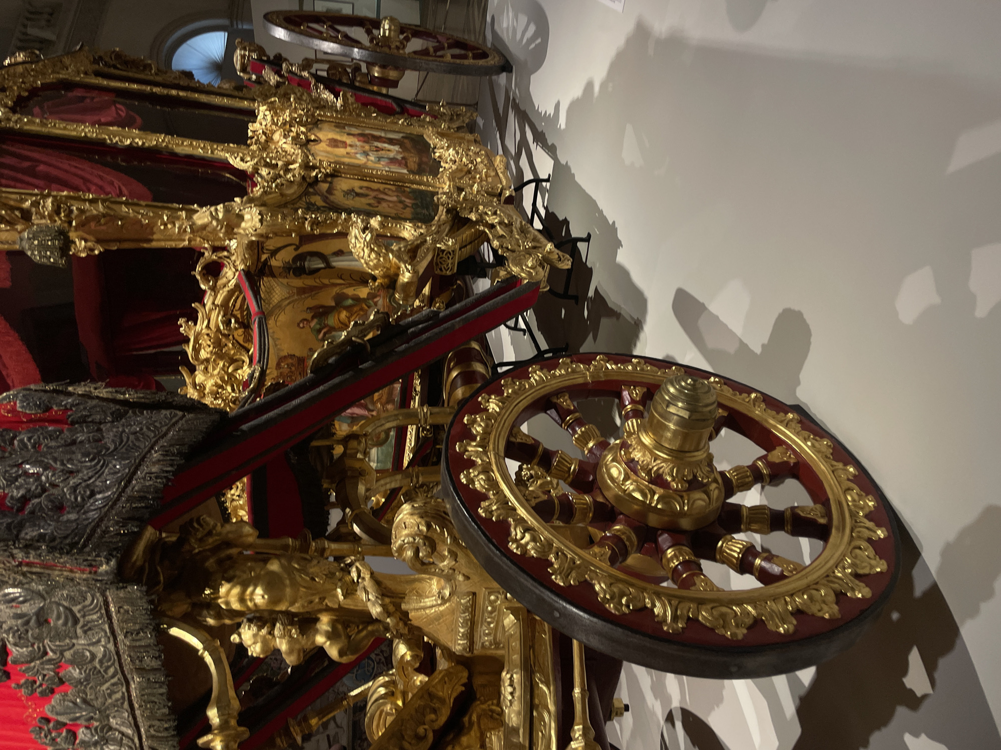 The image shows a section of an ornate golden carriage inside a building. The carriage features intricate carvings and decorations, with visible wheels adorned with gold accents. The carriage's construction includes a mix of red and gold elements. The scene is lit artificially, as evidenced by the shadows on the wall. The location is likely in Saint Petersburg, Russia, given the style and opulence often associated with Russian imperial artifacts.