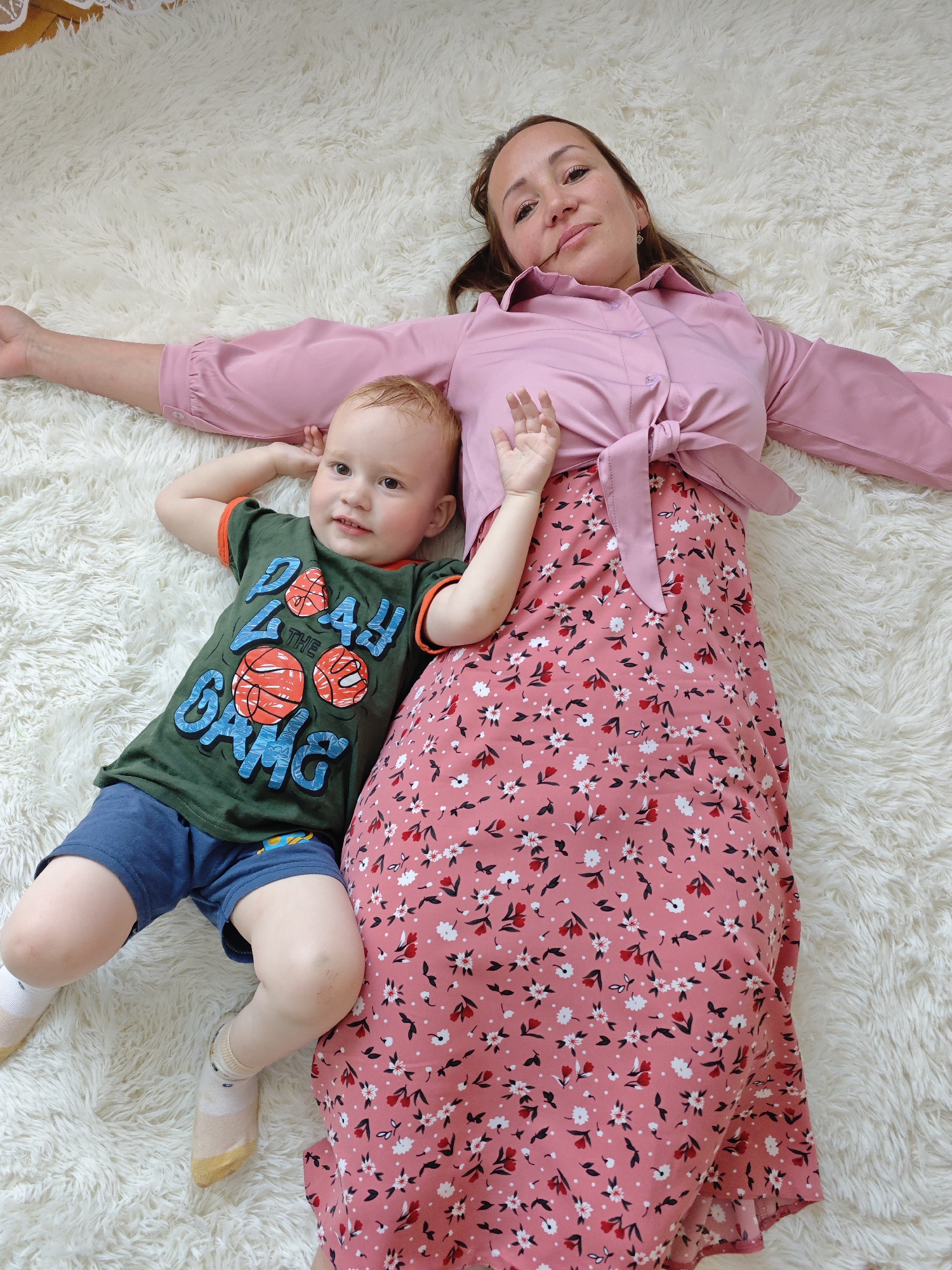 A woman and a young child are lying on a white, fluffy rug. The woman has medium-length brown hair and is wearing a pink button-down shirt tied at the waist and a long pink floral skirt. She is lying on her back with her arms extended to the sides. The child is lying on his back to the woman's left. He has red hair and is wearing a green t-shirt with a basketball design and blue shorts. The child's left arm is raised in the air. The location is indoors, presumably in a residence. Марковский, Russia is not verifiable in this image.