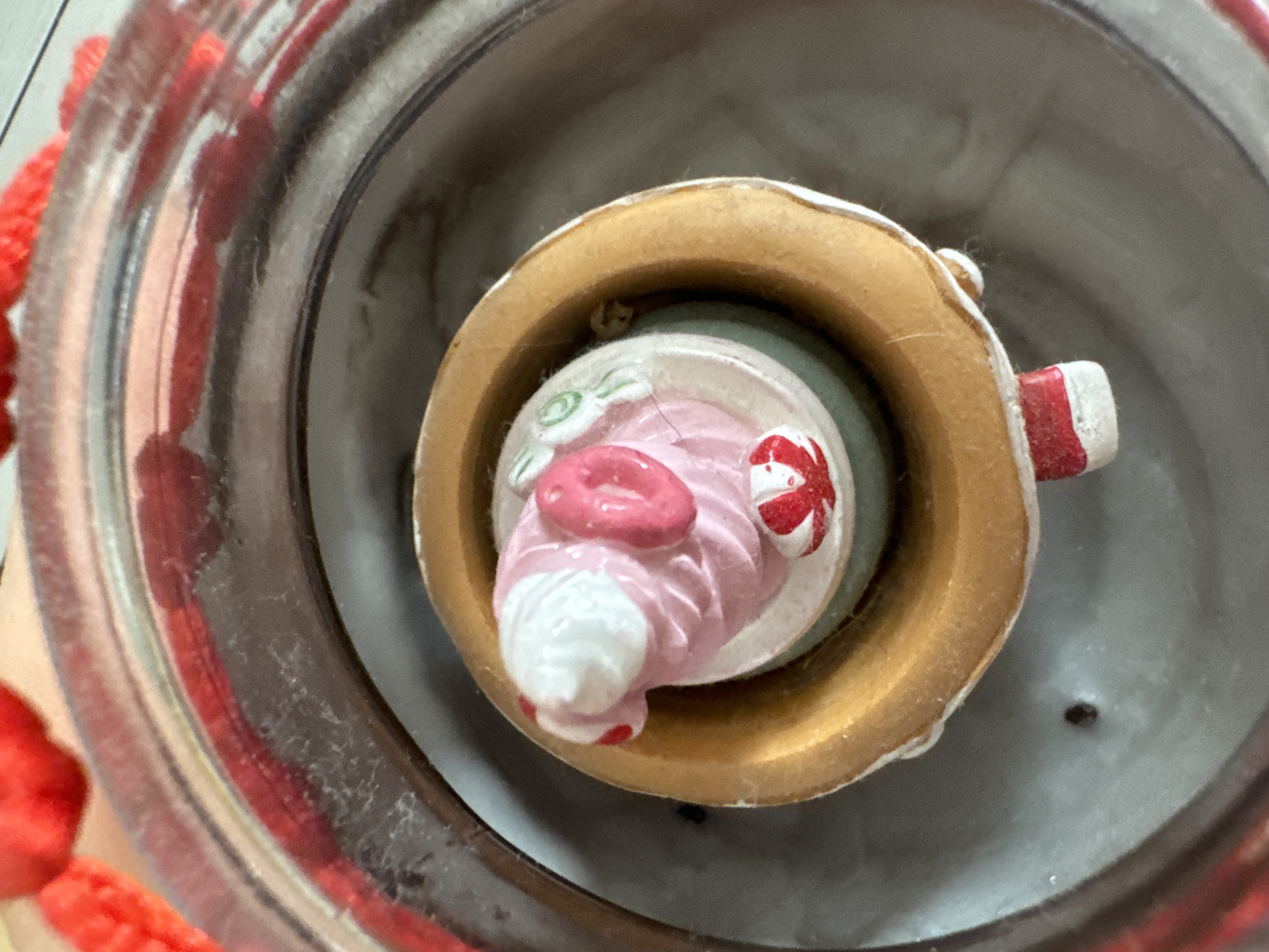 The image shows a close-up, top-down view of a small, decorative figurine inside a glass jar. The figurine appears to be a pink cupcake with white icing, adorned with red accents. It sits within a golden circular base, which is itself placed on a greenish layer. Red yarn or string is visible around the rim of the jar, suggesting it may be part of a larger craft or decoration. The interior of the glass jar is visible, showing a grey, slightly textured surface. The overall setting is unclear, but the objects are arranged in a static, decorative manner. There is no discernible indication of the city of Murcia, Spain, within the image itself.