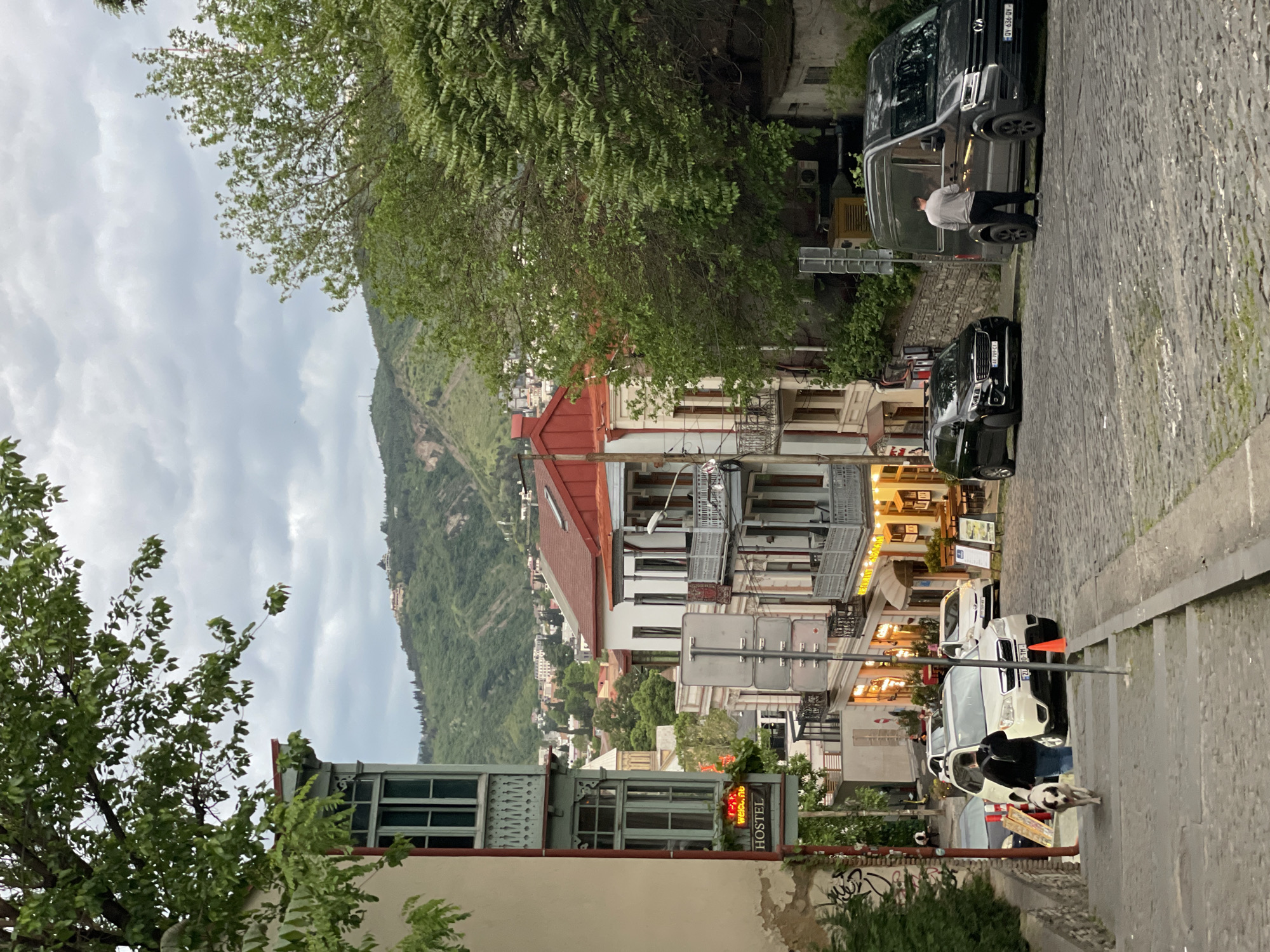 The image shows a street scene in Tbilisi, Georgia, looking downhill on a cobblestone street. Several buildings line the street, varying in height and architectural styles, some with red roofs and balconies. A hillside with green vegetation is visible in the background. A "HOSTEL" sign is noticeable on one of the buildings. A few parked vehicles are present, including black vans. A man stands near one of the vans. Further down the street, several parked cars are visible, along with what appears to be a person walking a dog. The sky is overcast.