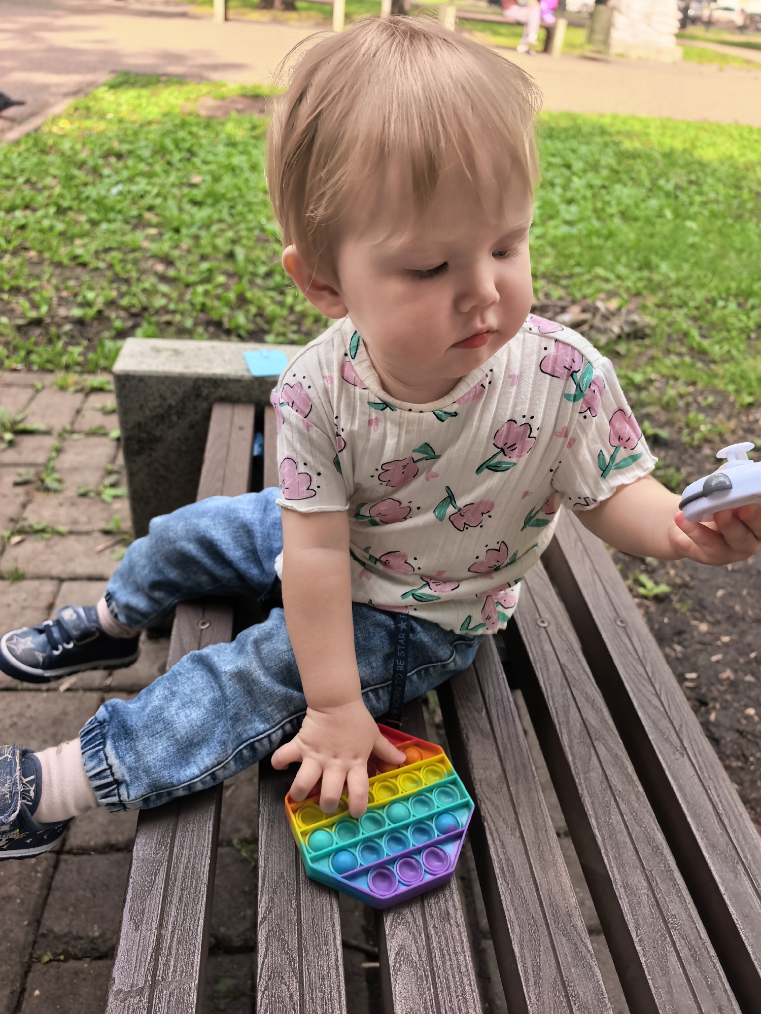 A young child with blond hair sits on a wooden park bench, playing with a rainbow-colored hexagonal fidget toy. The child is wearing a white t-shirt with a floral pattern, blue jeans, and dark blue sneakers with white star accents. The child's left hand is reaching for the fidget toy, while the right hand holds a white toy. The background shows green grass and trees, suggesting an outdoor park setting. There are no specific details in the image that indicate the location is the city of Sterlitamak, Russia.