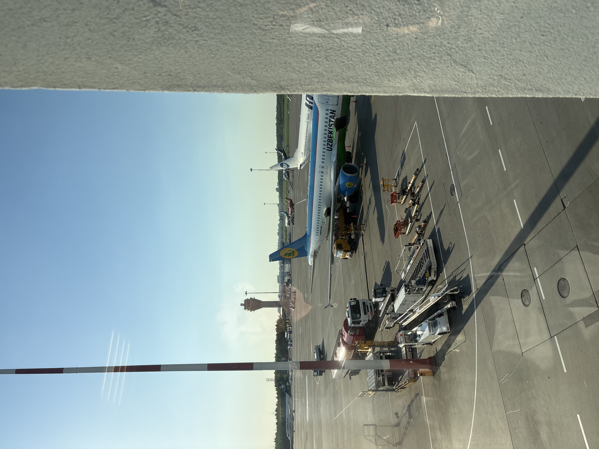 The image shows an airport scene, likely from a window within a terminal or adjacent building. Two Uzbekistan Airways airplanes are visible on the tarmac, being serviced by ground vehicles. There is a control tower visible in the background. The sky is clear and blue. The location is likely Pulkovo Airport in Saint Petersburg, Russia given the airline and the location.
