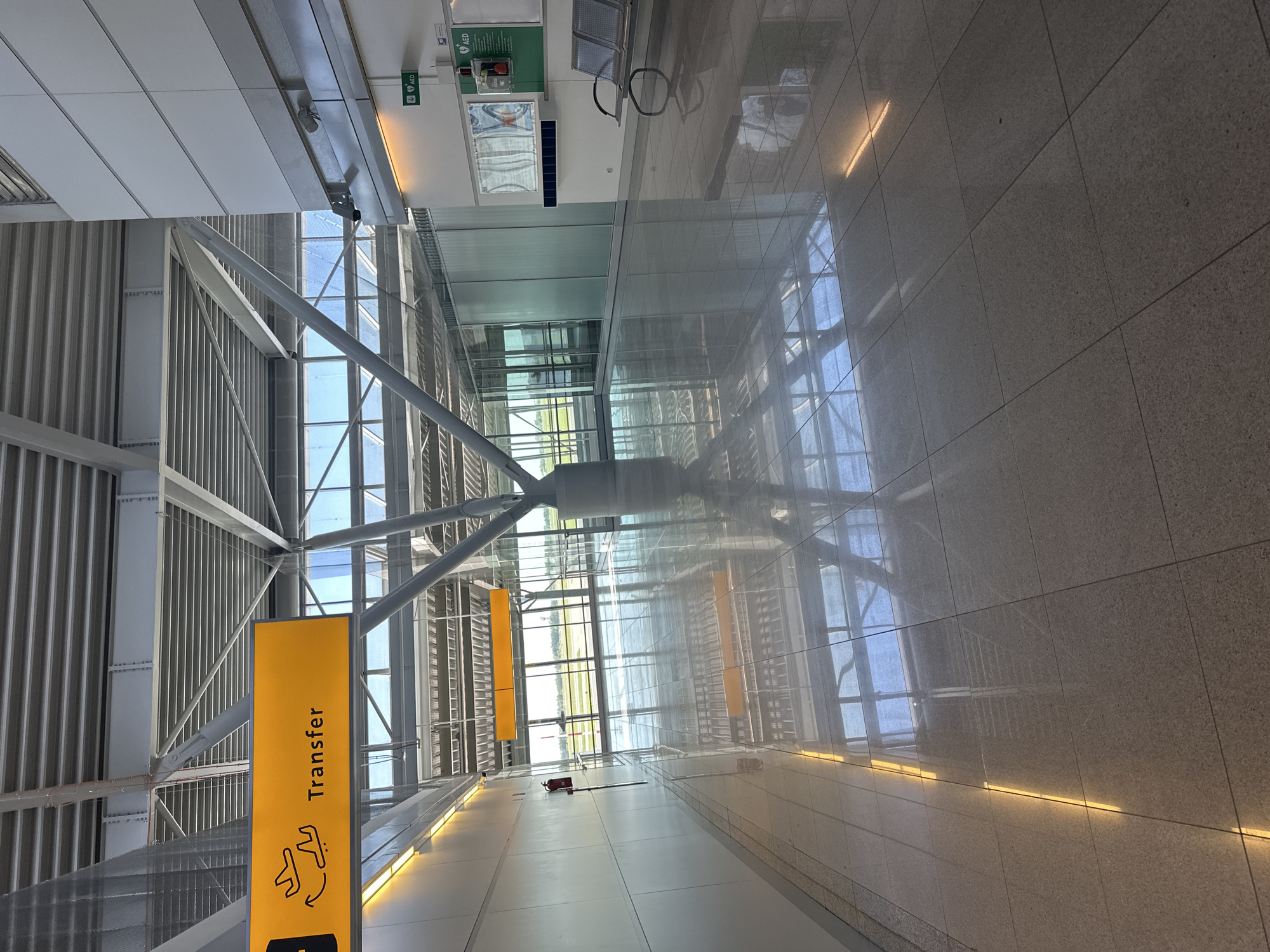 The image shows an interior view of an airport terminal with a long, reflective tiled floor. A yellow sign with the word "Transfer" and an airplane icon is visible overhead. The terminal has a modern industrial design with a high ceiling supported by exposed metal beams. Large windows offer a view of an outdoor area, likely an airfield. Seating areas and an AED station are present on the right side of the hallway. The scene is well-lit by natural light and internal lighting. The location is the Warsaw Chopin Airport.