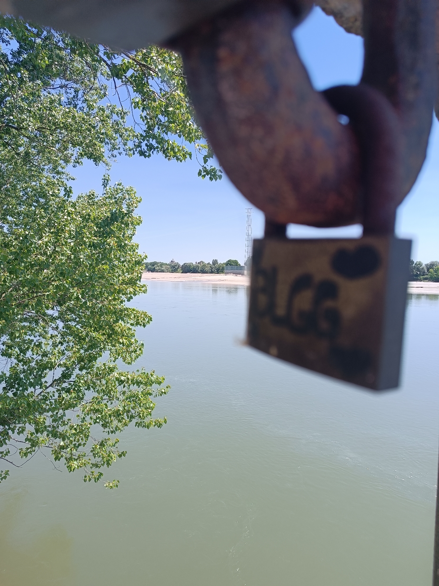 The image shows a river landscape in Viadana, Italy, viewed through a rusted metal chain with a love lock. The lock is out of focus and has faded letters "BLGG" with two heart shapes on it. A tree with green foliage occupies the left side of the frame. Across the river, there is a sandy bank and trees. An electrical tower is visible on the other side of the river. The sky is clear and blue. The river is pale green.