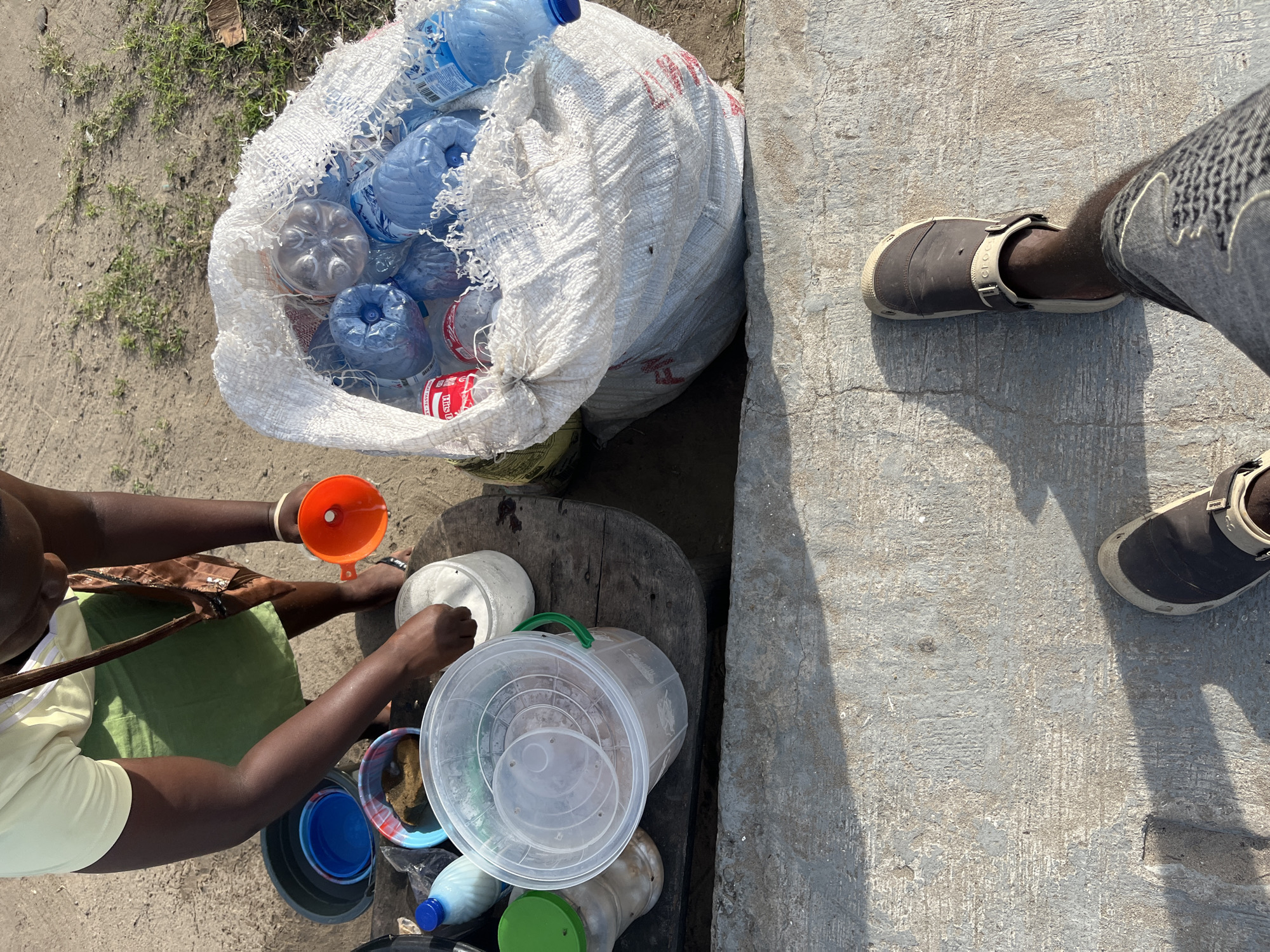 An individual is depicted pouring contents through an orange funnel into a white bucket, while a large white sack filled with numerous blue plastic water bottles is adjacent. This is done on a rough wooden table. Various other containers and bottles are situated on the table around the bucket. The setting appears to be outdoors, with a concrete or paved surface visible nearby, and patches of sandy ground with sparse vegetation in the background. Another individual is standing on the concrete ground, adjacent to the table, wearing gray jeans and a brown and beige shoe. The scene is in Badagry, Nigeria.