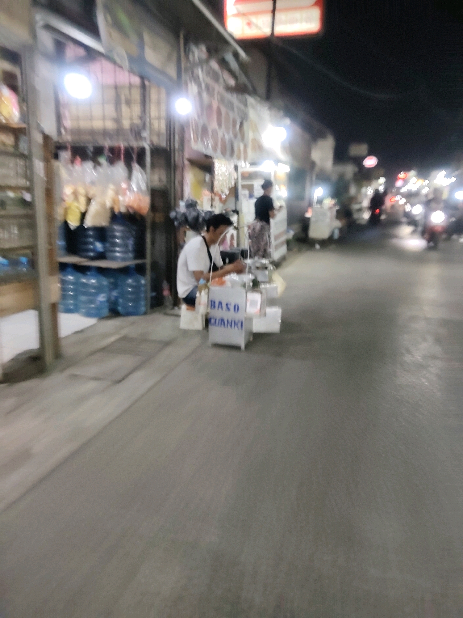 The image depicts a nighttime scene on a street in Mekarmukti, Indonesia. A vendor is operating a food cart, identified by the label "BASO CUANKI" on its side. The vendor, a person with dark hair wearing a white t-shirt, is behind the cart, appearing to be engaged in preparing or serving food. The cart is situated on a paved road, with a sidewalk or building frontage to the left. Several large blue water bottles are stacked on shelves to the left of the food cart, suggesting a retail or service area. In the background, other food carts and stalls are visible, along with illuminated signs and the headlights of passing motorcycles, indicating a busy street market or food stall area at night. The overall impression is one of a typical urban street scene with commercial activity occurring after dark.