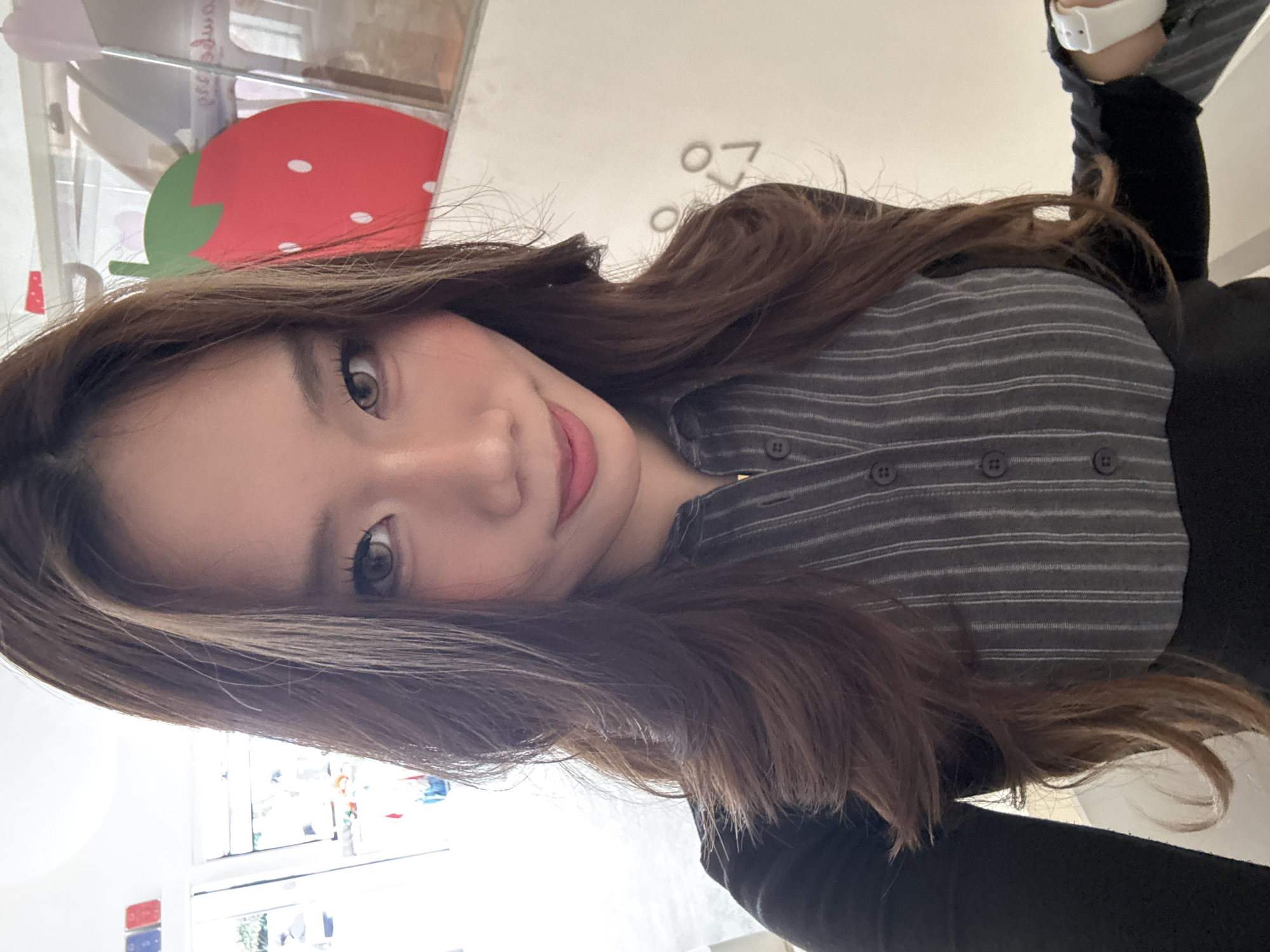 A woman is positioned at an angle in the foreground, appearing to be taking a selfie. She is wearing a striped button-up shirt over a black top. She has long, brown hair and is wearing makeup. In the background, there is a blurry, indistinct scene that includes a cutout of a strawberry, a white surface with indiscernible markings, and a window. The location, as stated, is in 江北城街道, China.