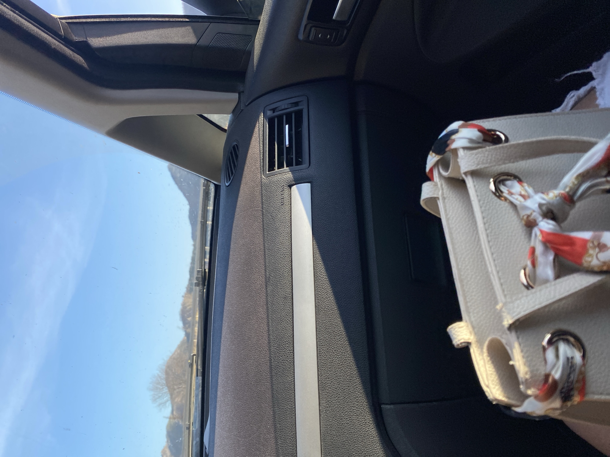 The image depicts the interior of a vehicle, likely a car, with a view through the windshield to the exterior. The dashboard is visible, featuring a ventilation grill and a silver trim piece. A handbag, adorned with a patterned scarf, is partially visible on the right side of the frame. The outdoor scene shows a clear, bright sky and a landscape with sparse trees and low hills or mountains in the distance. Given the prompt's reference, the location is purportedly Golubovići, Montenegro.