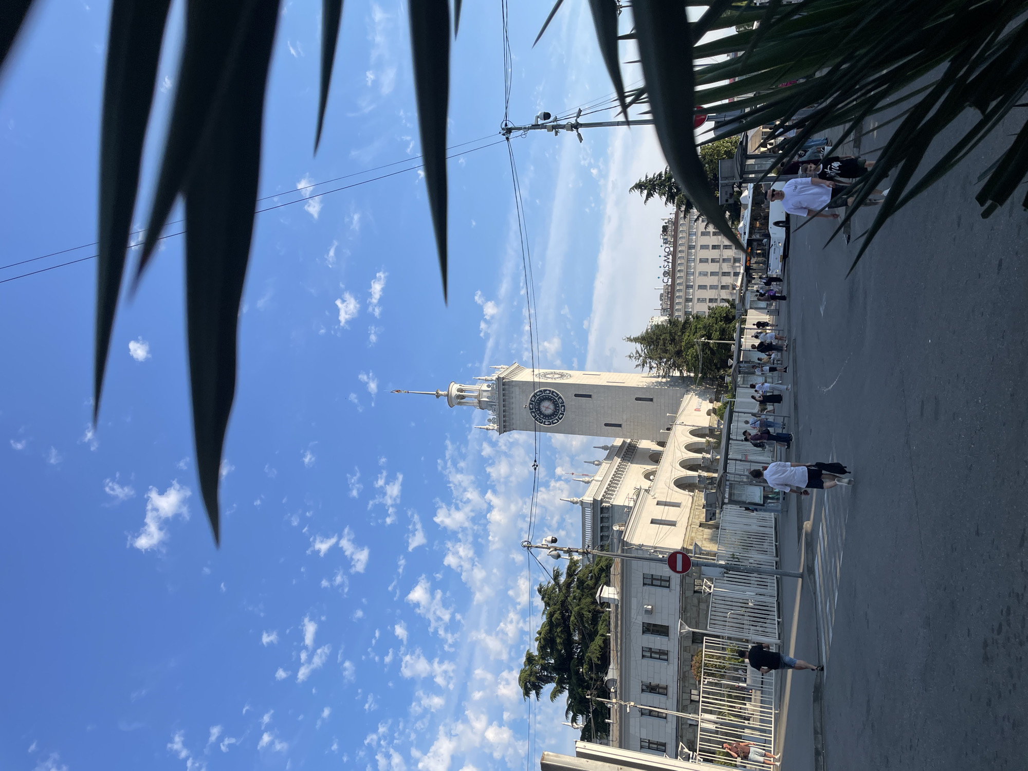 This media file depicts a daytime scene in Sochi, Russia, likely a public square or street with a significant building. The foreground is partially obscured by the fronds of a palm-like plant, creating a natural frame.
The dominant architectural feature is a tall, white clock tower with a circular clock face featuring Roman numerals and ornate hands. Adjacent to the tower is a long, multi-story building with arched windows and decorative elements, suggesting a historical or official structure. The sky is a bright blue, dotted with scattered white clouds, indicating pleasant weather.
Several people are visible on a paved area, suggesting a public space. Some are walking, while others appear to be standing or gathered. Their attire, including t-shirts and shorts, is casual and appropriate for warm weather. A bus is parked on the side of the road, and there are power lines crisscrossing the sky, typical of an urban environment. A red and white circular sign with a horizontal bar, indicating "no entry" or a similar prohibition, is mounted on a pole near the building.
In the background, another large, light-colored building with numerous windows is visible, possibly a hotel or residential complex. Green trees, including some tall conifers, are interspersed throughout the scene, adding to the natural setting.
The overall impression is a bustling public area on a sunny day, with a mix of architectural grandeur and everyday activity. The time of day appears to be late morning or early afternoon, judging by the sunlight and shadows.