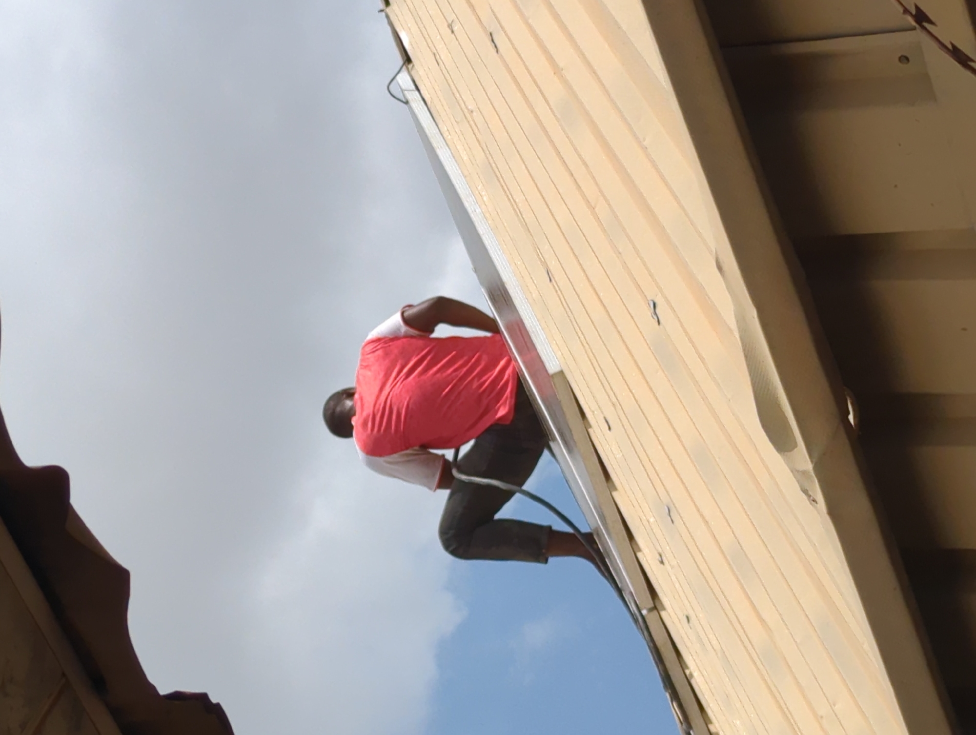 An upward angle captures a person on a ladder against a light-colored wooden structure, with a cloudy sky in the background. The individual wears a pink t-shirt over a white long-sleeve shirt and dark jeans. They stand on the ladder using one foot while holding a black hose. The setting appears to be Sabon Tasha, Nigeria.
