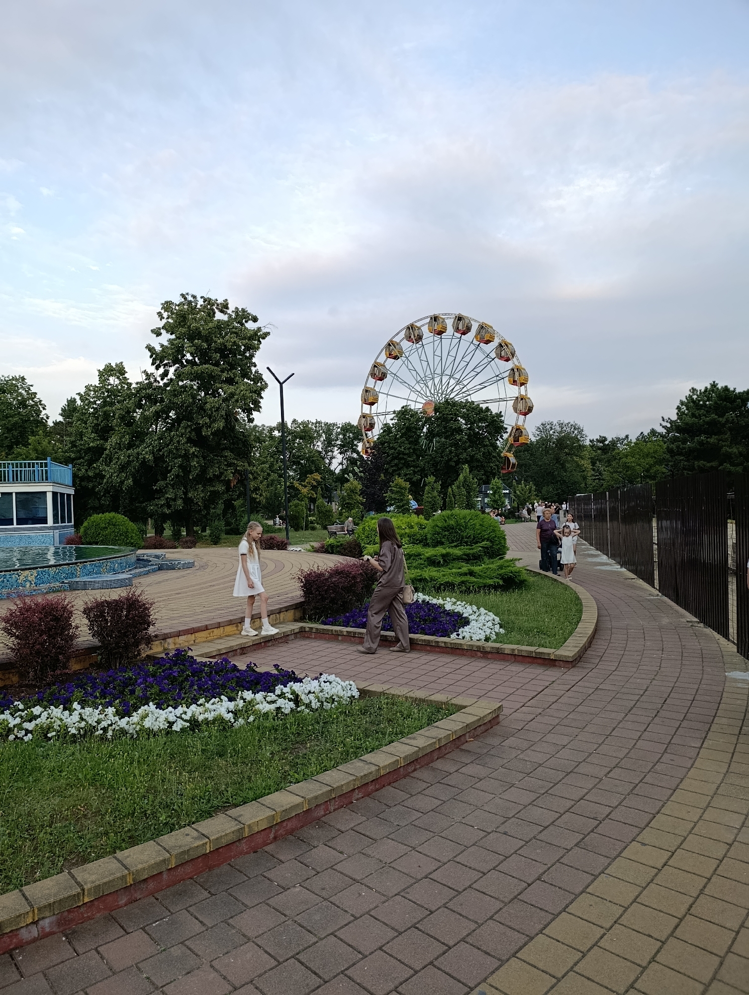 The image captures a park scene in Maykop, Russia, featuring a large Ferris wheel in the background, partially obscured by trees. In the foreground, a paved walkway curves through the park. Two young girls are walking on the walkway, one in a white dress and the other in a brown outfit. There are flower beds with purple and white flowers lining the walkway. To the left, a building with a blue roof and a water feature is visible. The sky is overcast, indicating a cloudy day. The overall atmosphere is calm and recreational.