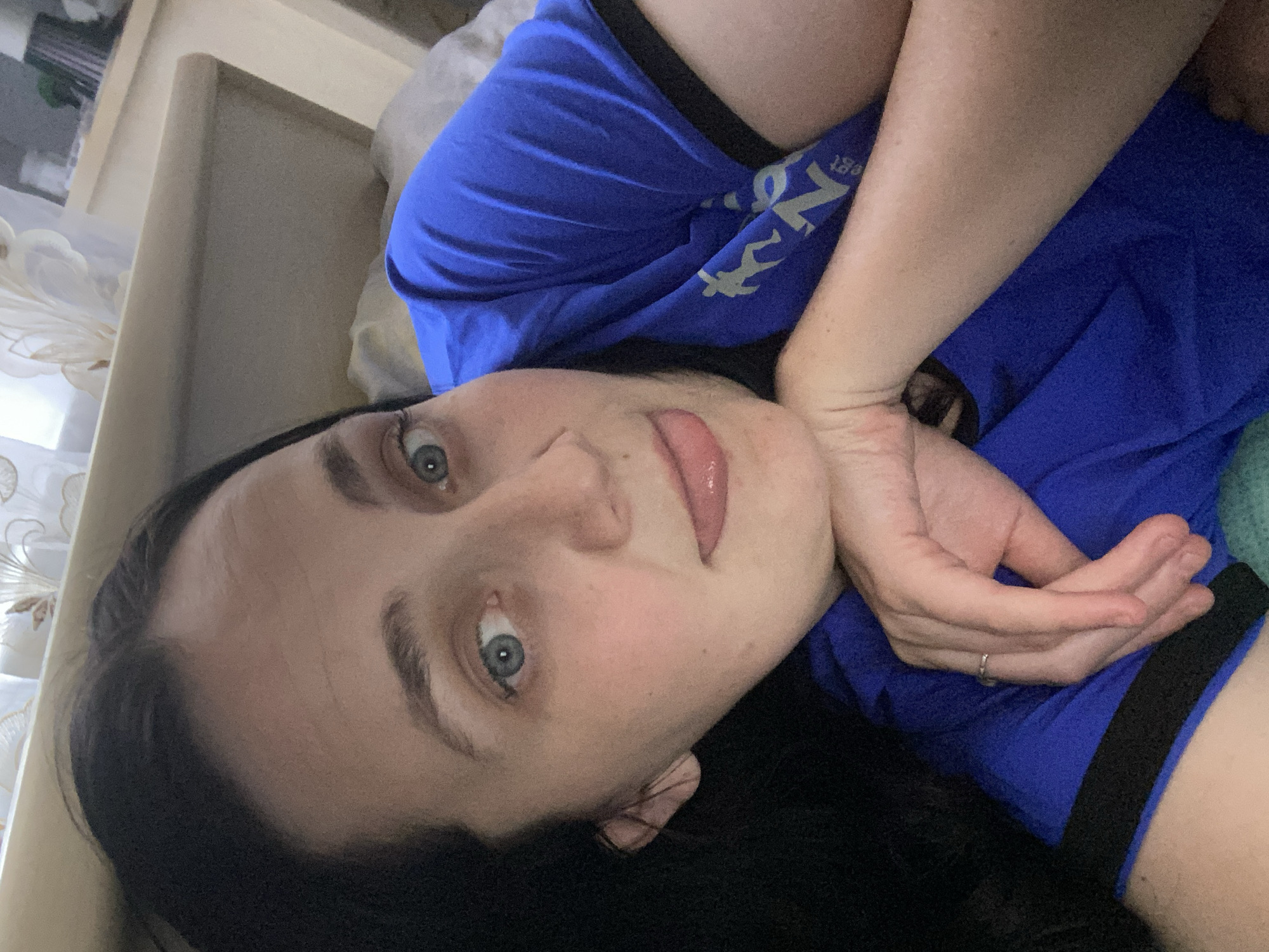 The image is a close-up, selfie-style shot of a woman lying down, with her head resting on her arm. She has dark hair and blue eyes, and she is wearing a blue t-shirt. Her tongue is slightly sticking out. The background appears to be a bedroom, with a light-colored headboard and curtains visible. The lighting is soft and diffused. The woman is the sole subject in the image. There are no other people or animals present. The environment is domestic and appears to be indoors. The image is taken from a high angle, looking down at the woman.