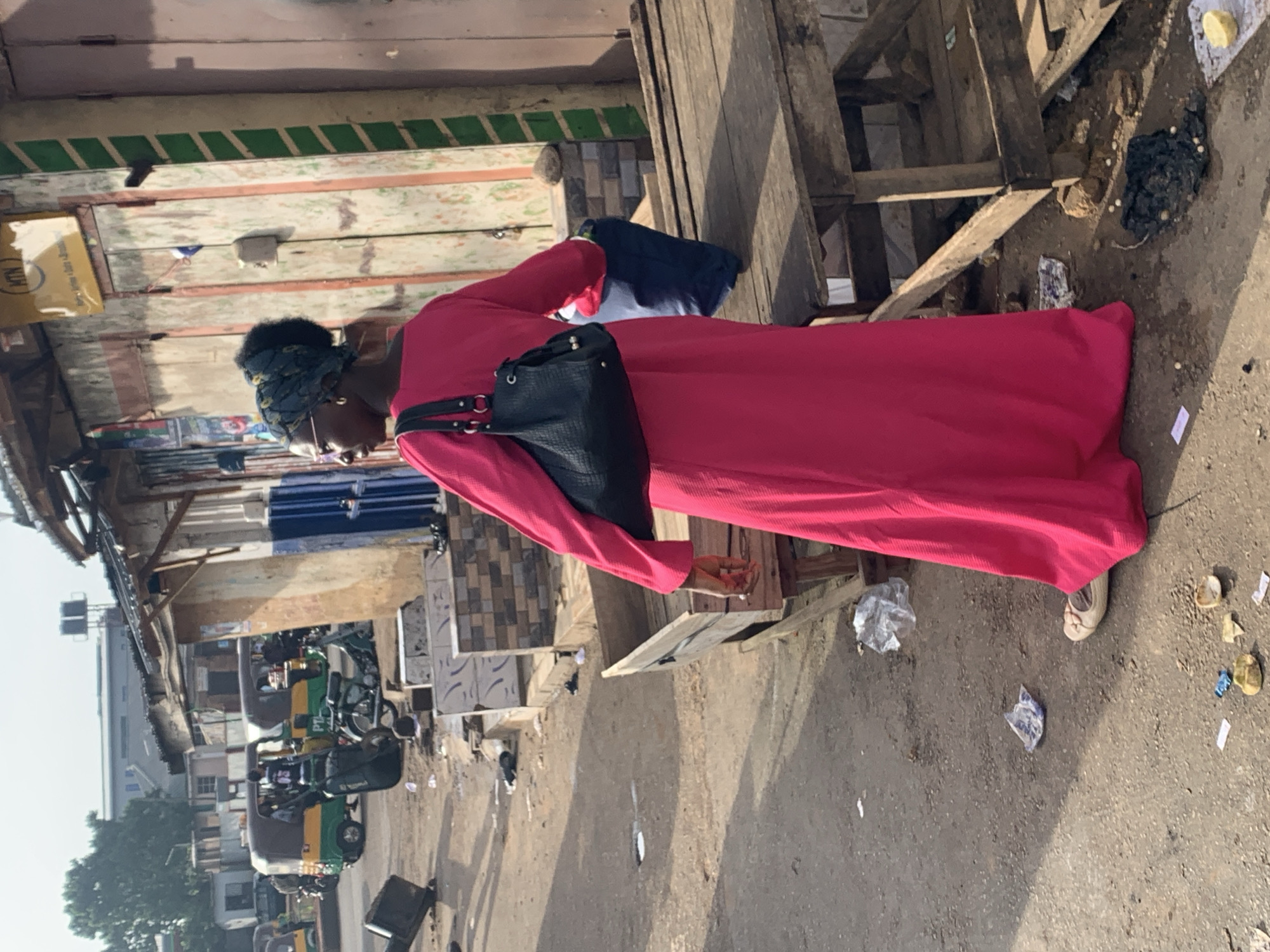 In Gura Top, Nigeria, a woman stands on a dirt road, facing away from the camera. She is wearing a bright pink, floor-length dress and a dark headscarf. A black handbag with a textured surface hangs over her left shoulder. The background shows a street scene with several open-fronted market stalls. One stall has a yellow sign with the "MTN" logo. In the distance, several auto-rickshaws, a common mode of transport in Nigeria, are parked or driving on the road. The ground is littered with various items, including plastic bags, wrappers, and some scattered objects. The scene appears to be a bustling market area.