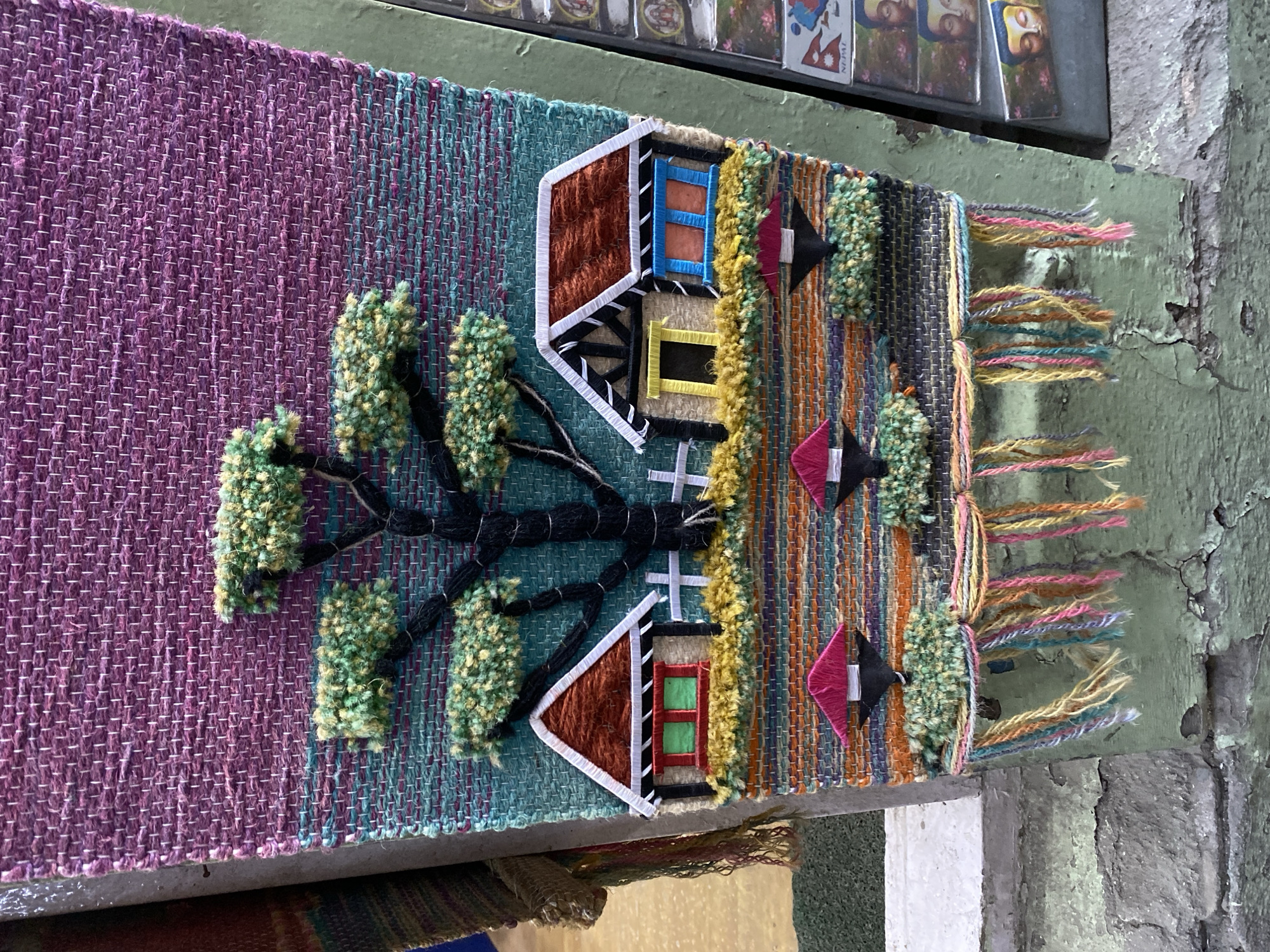 The image shows a close-up view of a colorful, woven tapestry depicting a rural scene with houses, a tree, and a fence. The tapestry is hanging on a green, weathered pillar, possibly in an outdoor market or street setting in Kathmandu Metropolitan City, Nepal. The background shows hints of other items, suggesting a commercial area. The tapestry itself is intricately designed with various textures and colors, featuring a large tree with green foliage, two small houses with red roofs and colorful windows, and a few smaller structures with pink roofs. The bottom of the tapestry has tassels of various colors. To the right of the tapestry, a stack of small souvenir cards with images of Buddha and text that reads "NEPAL" are visible, further confirming the location. The ground surface below the pillar appears to be a rough, textured surface, possibly concrete or stone, with some wear and tear.