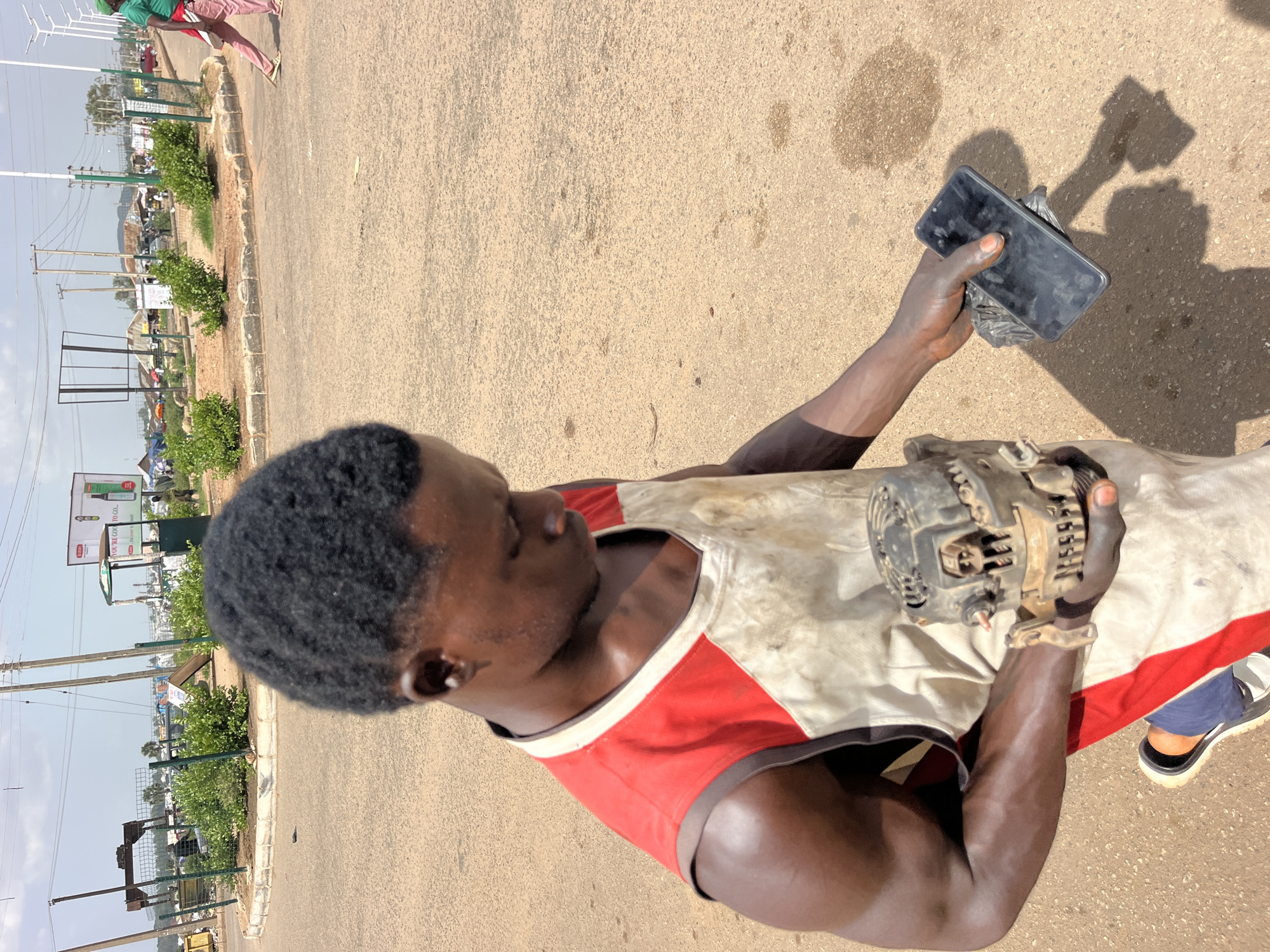 A young Black male is standing in a sandy, asphalt-covered area that appears to be a road or a roundabout in an urban environment. He is wearing a white and red sleeveless garment that is stained with grease and dirt, suggesting manual labor. His hair is short and dark. In his right hand, he holds a black smartphone. In his left hand, he holds a dirty, metallic object that resembles a car alternator. His gaze is directed downwards, focused on the items in his hands. The background shows a slightly hazy sky with scattered clouds, power lines, billboards, and some structures that suggest a busy, possibly developing, area. There are also several green bushes and a few other people in the distance, one of whom is walking away from the camera. The ground is uneven with some dark spots, possibly oil stains.