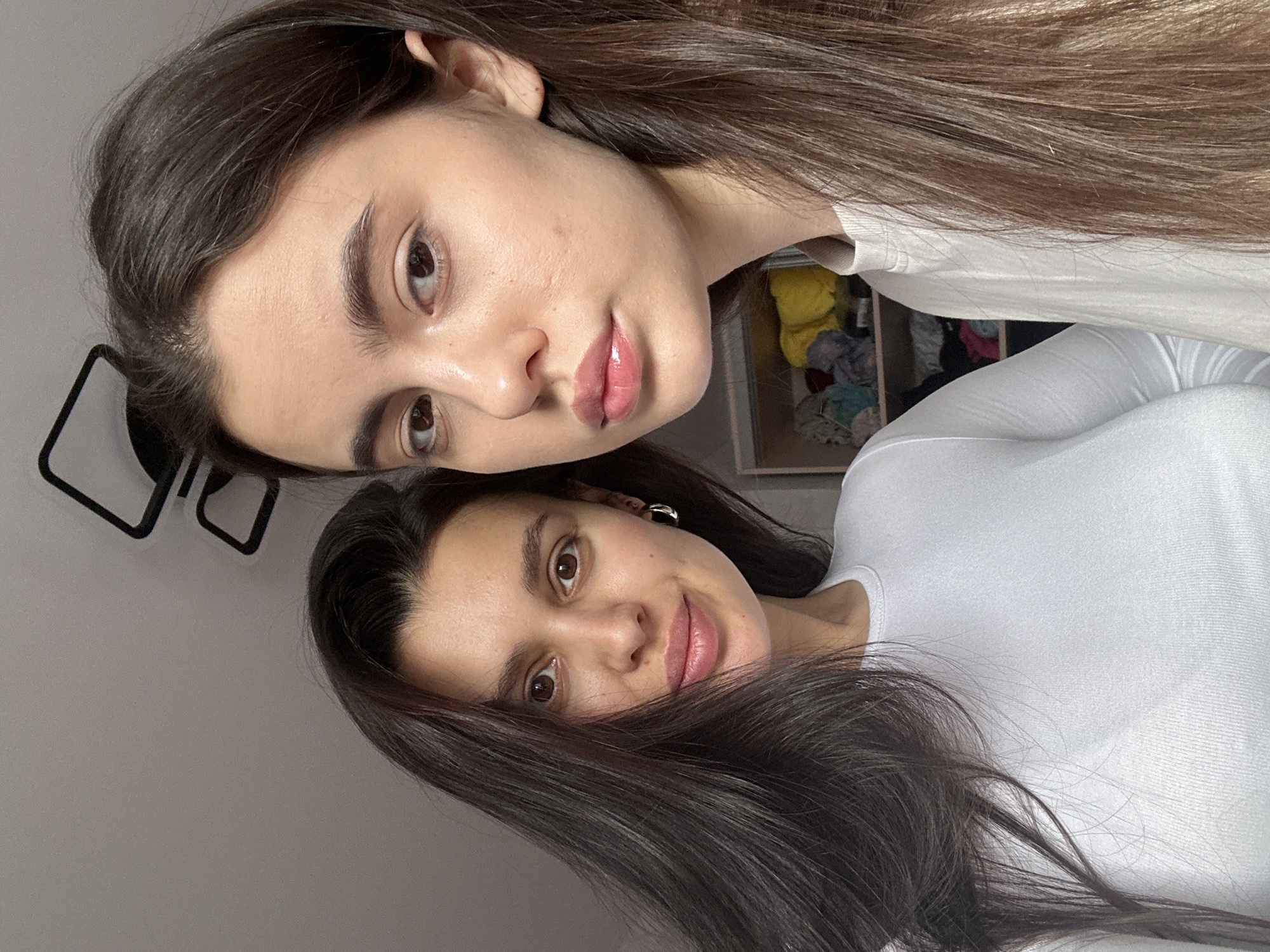 The image displays two young women posing for a casual portrait indoors, rotated 90 degrees counter-clockwise. Both subjects have long, dark brown hair and are wearing plain, light-colored tops, likely white or off-white.
The woman in the foreground is positioned closer to the viewer, looking directly at the camera with a neutral to slightly pleasant expression, her lips appearing glossed. Her hair flows past her shoulders. The second woman is partially visible behind and above the first, also facing the camera with a neutral expression. Her hair frames her face.
The background indicates an indoor setting, possibly a bedroom or living space. A black, abstract, angular object, possibly a lamp or decorative item, is partially visible in the top-left corner. Behind the upper woman, a shelf or cubby is discernible, holding several neatly folded items of clothing or fabric in various colors. The lighting is soft and even, suggesting either diffuse natural light or well-distributed artificial light. There are no specific visual cues within the image to identify the time of day or weather. No visible text is present. The setting is a typical casual indoor environment in Chaikovsky, Russia.
