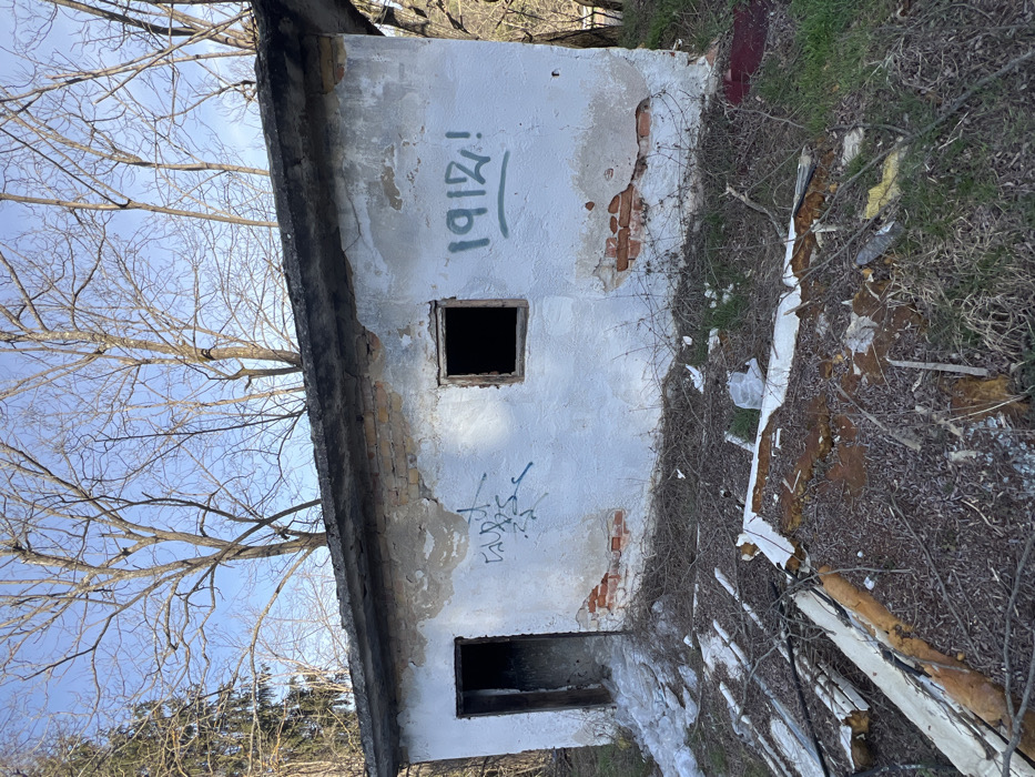 An abandoned, single-story, rectangular structure with heavily degraded white plaster walls, revealing red brickwork, stands in the city of Stoianovo, Bulgaria. The building features an open doorway on the left and an empty window opening on the right, both devoid of frames or coverings, leading to dark interiors. Blue spray-painted graffiti is visible between the door and window, and "1912!" in green spray paint is present to the right of the window. The structure has a dark, flat concrete roof. The surrounding ground is covered with dry brush, bare earth, sparse green vegetation, and scattered debris including white panels with exposed insulation. Patches of snow are present near the doorway. Deciduous trees with bare branches are visible in the background against a clear blue sky. No individuals or active elements are present in the scene.