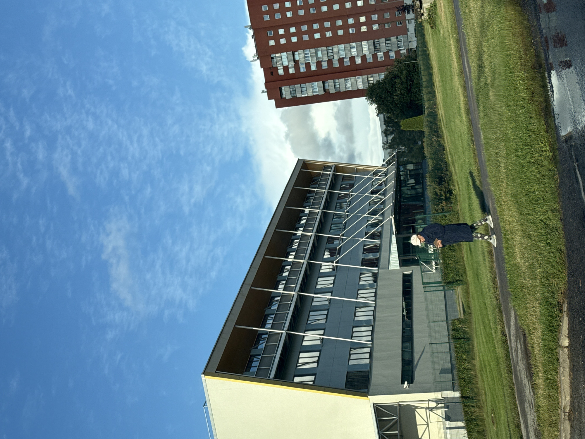 An outdoor, eye-level, medium shot captures a sunny day in Tallinn, Estonia. The scene features a light-colored, modern building with a yellow accent on the left, and a red, multi-story apartment building on the right. A person, wearing a dark coat, a light-colored hat, and patterned leggings, walks on a paved path in front of the light-colored building, holding a mobile device. The ground is covered in green grass, and a puddle reflects the sky and buildings. The sky is blue with scattered white clouds.