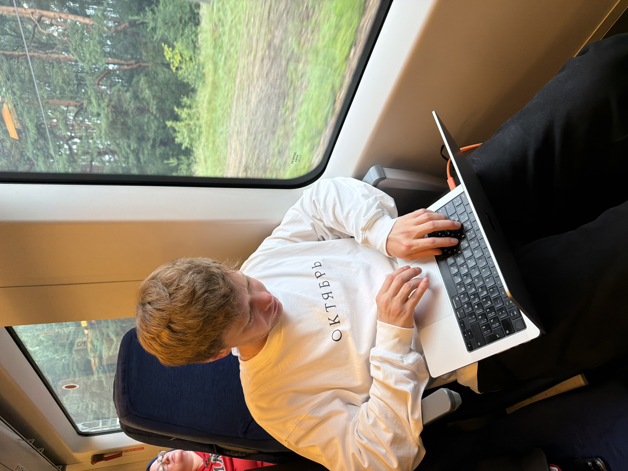 A young person with short, light brown hair sits in a train compartment, looking down at a laptop. They are wearing a white long-sleeved shirt with black text that reads "ОКТЯБРЬ" (October in Russian) across the chest, and dark pants. The laptop, a silver MacBook, is open on their lap, and their hands are on the keyboard. The train compartment appears to be moving, as evidenced by the blurred scenery of trees and grass visible through the window. In the background, another passenger wearing glasses and a red hoodie can be seen. The scene takes place inside a train traveling through a wooded area, likely in or near Nazar'evo, Russia, given the text on the shirt and the landscape.