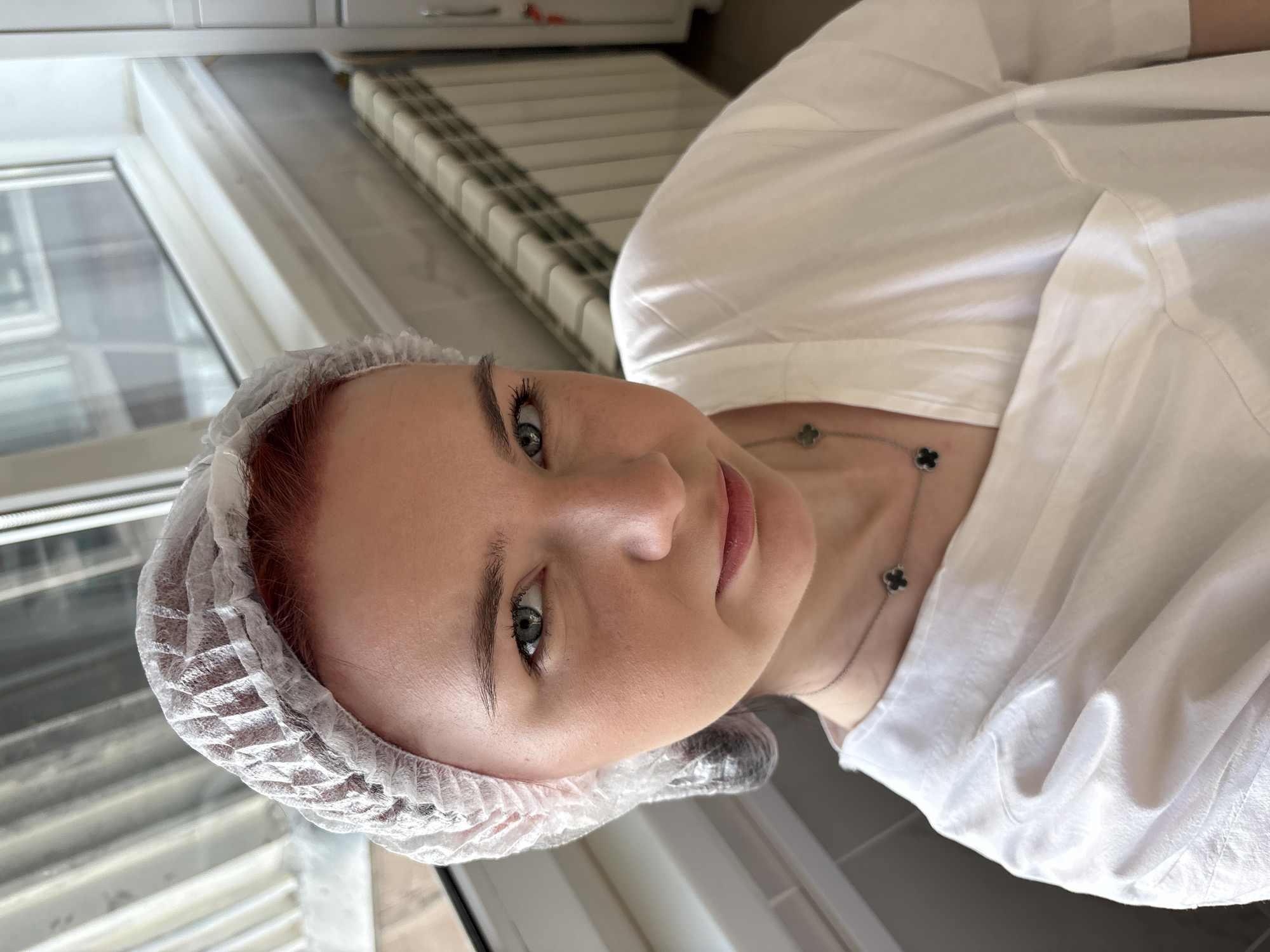 A young woman with red hair, wearing a white disposable hairnet and a white V-neck top, looks directly at the camera. She has light blue eyes and is wearing a silver necklace with four-leaf clover pendants. The background shows a window with blinds, a radiator, and a grey tiled wall, suggesting an indoor environment, possibly a medical or salon setting. The image is a close-up selfie, taken from a slightly high angle. There are no specific landmarks visible that would indicate a precise location within Saint Petersburg, Russia.