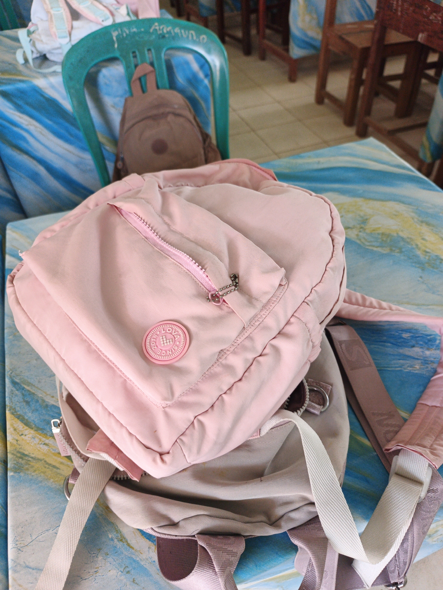 A close-up shot shows two pink backpacks on a table. The backpack in the foreground is a lighter shade of pink with a prominent circular emblem and a zippered pocket. The second backpack, partially obscured beneath the first, appears to be a darker shade of pink. In the background, a teal plastic chair is visible with a smaller, brown backpack slung over its back. The chair has writing on it that appears to be "fina. Anggun. O". The scene suggests a casual setting, possibly a school or community area, in Tanjung Setia, Indonesia. The table's surface has a colorful, marbled pattern of blues and yellows. The overall lighting is soft and diffused.