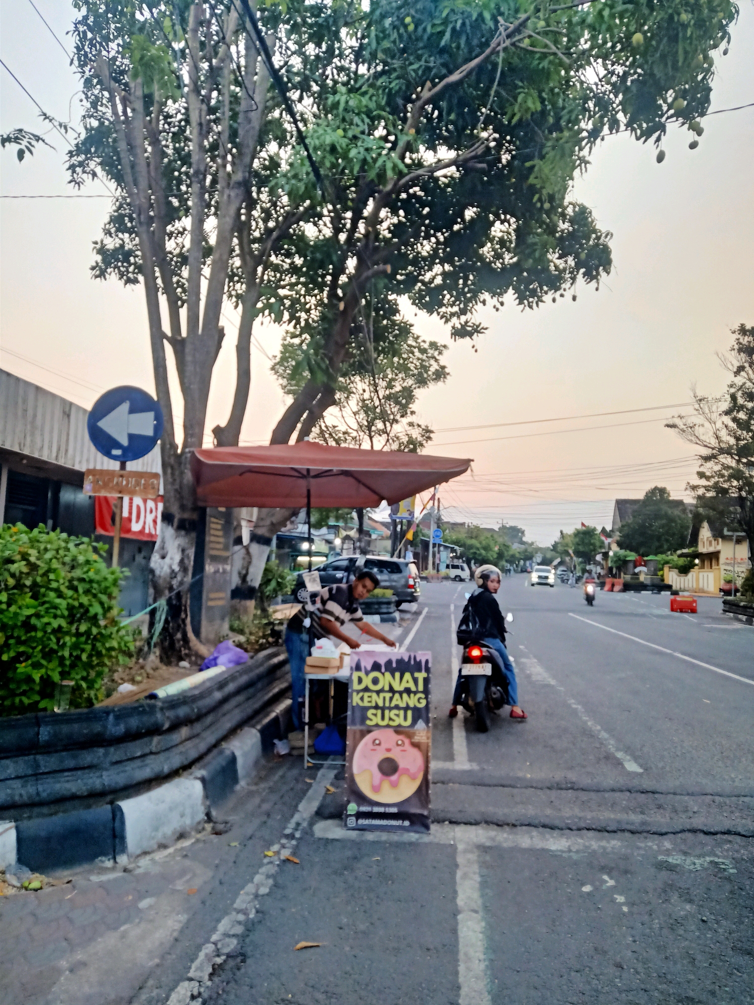 The image depicts a street scene in Klaten, Indonesia, during what appears to be late afternoon or early evening, as indicated by the soft, warm light and the hazy sky. A paved road runs horizontally across the frame, with a sidewalk and a grassy area to its left.
On the sidewalk, a food stall is set up, featuring an umbrella providing shade over a table where a vendor is preparing food. A prominent sign with an illustration of a donut and text in Indonesian, "DONAT KENTANG SUSU" (Potato Milk Donut), suggests the stall sells donuts. A person on a motorcycle, wearing a helmet and dark clothing, is parked near the stall, facing away from the camera.
Further down the road, vehicles, including cars and motorcycles, are visible, suggesting moderate traffic. Buildings line the street, with some architectural details suggesting a residential or commercial area. Lush green trees are scattered throughout the scene, with one large tree with multiple branches dominating the left side of the image. A blue traffic sign with a left-pointing arrow is also visible. The overall impression is that of a typical street scene in an Indonesian city, likely a place where street food vendors operate.