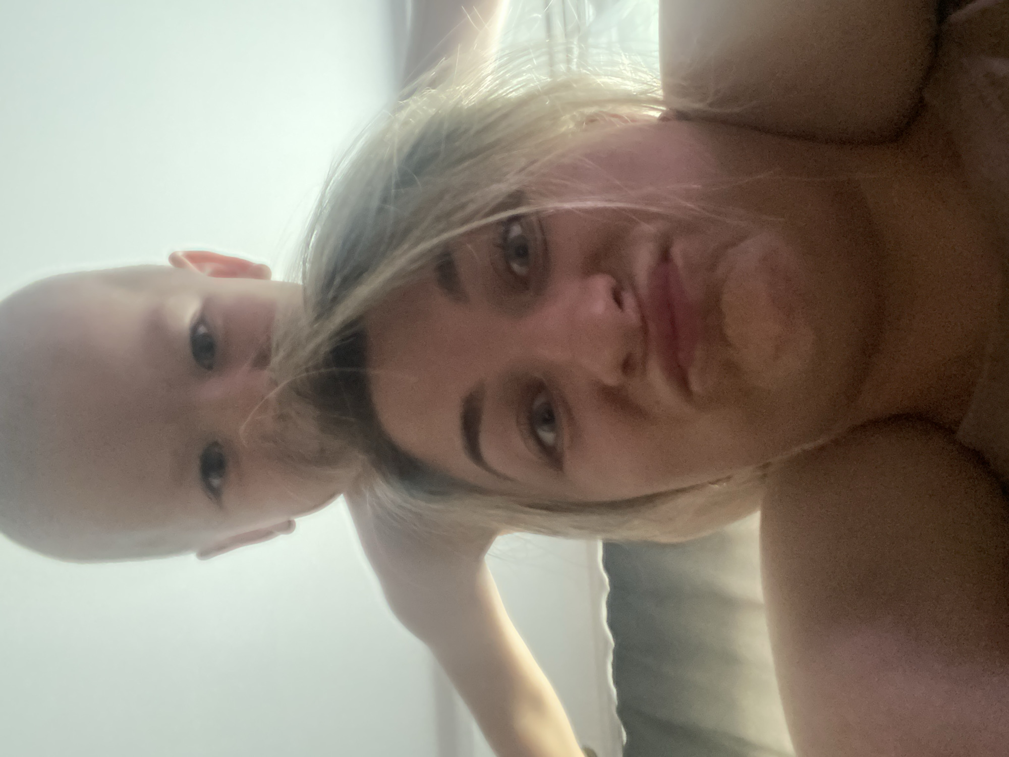 The image shows a low-angle, close-up, selfie-style photograph of a woman and a baby. The woman is in the foreground, her face is visible and she is making a pouting expression. Her hair is blonde and messy. The baby is positioned behind her head, with its face also visible, looking towards the camera. The background appears to be an indoor setting with bright lighting, possibly a room with a ceiling and a curtain or wall in the distance. The woman is wearing a light-colored top, and the baby is bald. The photograph was taken in Prokopyevsk, Russia.