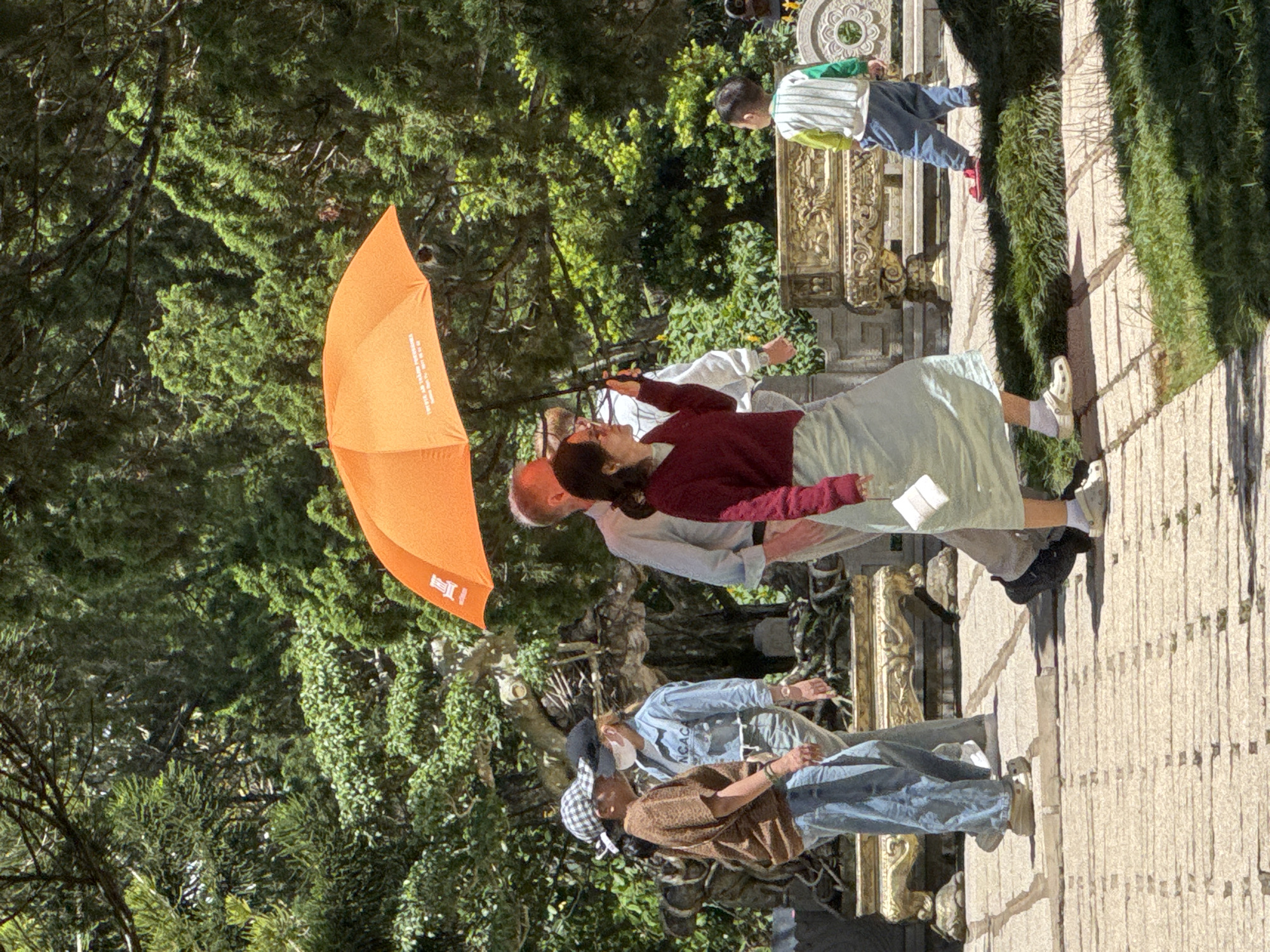 In Da Lat, Vietnam, a group of people walk through a landscaped area. An orange umbrella shields a woman in a red sweater and light blue skirt from the sun. She holds a white tissue in her left hand. Beside her, a man in a light blue shirt walks with his right hand near his pocket. Behind them, two women and a young boy are also walking. One woman wears a brown patterned shirt and a checkered hat, while the other wears a denim shirt and a black cap with a face mask. The boy, facing away from the camera, wears a striped shirt and blue pants. The ground is paved with light-colored stones, and lush green trees and ornate golden benches are visible in the background.