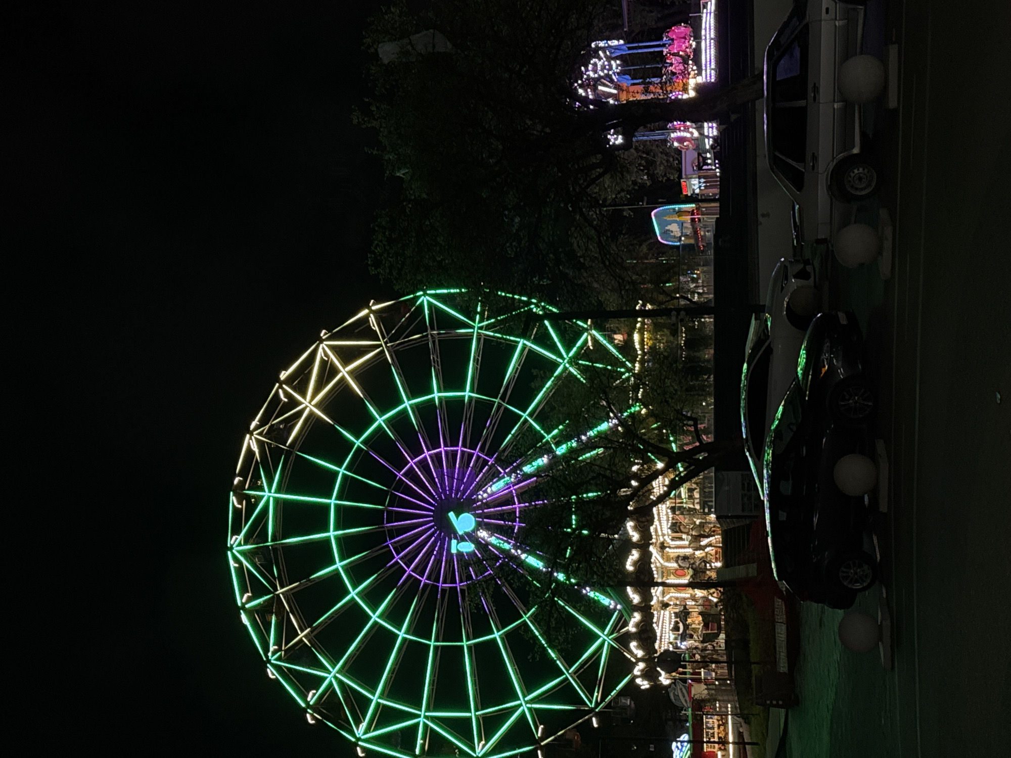 At night, a brightly lit Ferris wheel with green, purple, and yellow neon lights dominates the left side of the frame. In the background, other amusement park rides are visible, also illuminated. To the right, several cars, including what appears to be a black sedan and a white vehicle, are parked. White spherical lights are placed along the edge of what seems to be a sidewalk or walkway. The location appears to be in Novorossiysk, Russia, based on the identifiable architectural details and the type of amusement park present.