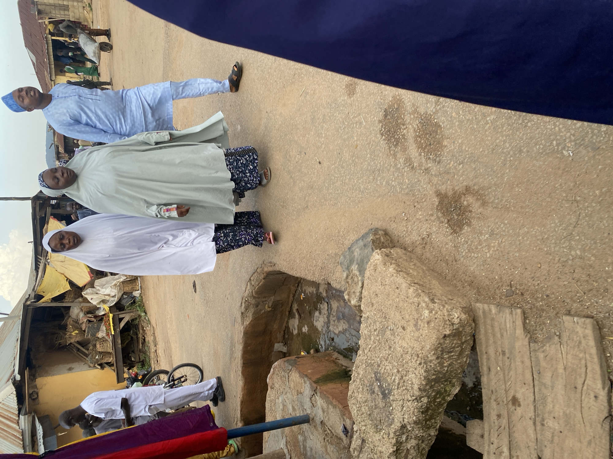 This image captures a street scene in Jos, Nigeria, likely in a market area. The foreground features a rough, textured cement structure bordering a shallow, water-filled ditch or drain, with two weathered wooden planks placed across it. Behind this, a paved road extends into the background, populated by several individuals.
In the middle ground, three women, all wearing Islamic attire (hijabs or abayas), are walking. The woman on the left is in a white abaya, the one in the center is in a light grey abaya, and partially visible on the right is another figure in a dark blue garment, likely also a woman. A man in a light blue traditional outfit and a blue cap is walking alongside the women, facing right and speaking. Another man in a white outfit, with a white cap, is seen on the left, near a parked motorcycle with a child's bicycle attached to its rear.
The background reveals a bustling market environment with stalls constructed from corrugated iron and fabric awnings, displaying various goods including baskets. The overall setting suggests a lively, everyday urban scene in Jos.