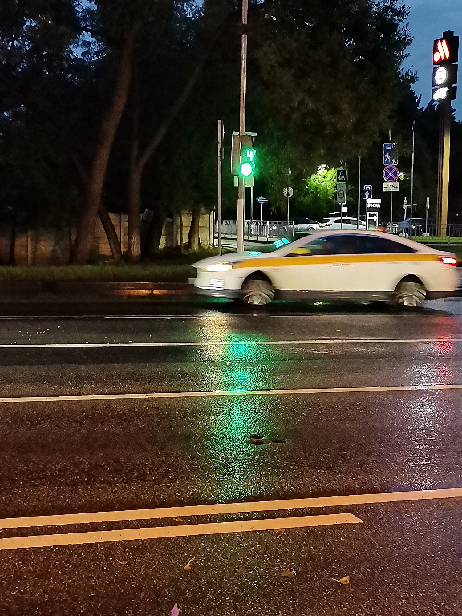 The image depicts a street scene at night, likely in Krasnogorsk, Russia, given the visual cues. A white car with a yellow stripe is driving on a wet asphalt road, reflecting the green light of a traffic signal. The road has double yellow lines painted on it, indicating a dividing line. In the background, trees line the street, and several traffic signs are visible, including a pedestrian crossing sign and a sign with a red circle and a diagonal line, likely prohibiting something. A traffic light is also visible, showing a green signal. The overall impression is of a city street at night, possibly after rain, with a car in motion.