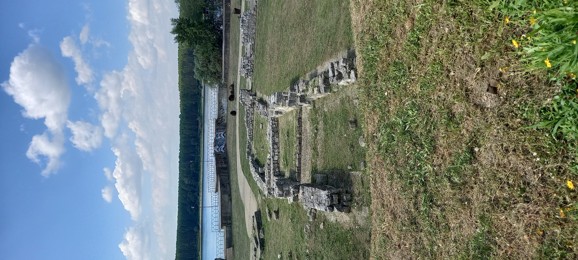 This is a medium-shot, vertical, outdoor, daytime photograph taken in Silistra, Bulgaria. The image depicts the ruins of an ancient stone structure in the foreground. The ruins consist of low stone walls outlining rectangular rooms and pathways. The ground is covered in green and dry grass, with scattered rocks and some yellow wildflowers.
In the middle ground, there is a paved walkway leading towards a concrete embankment. On the embankment, there are several black, cylindrical objects, possibly barrels or large metal containers. The embankment is lined with a blue metal railing. Graffiti is visible on the concrete wall of the embankment.
The background features a wide, blue river with a dense line of green trees along the opposite bank. The sky above is blue with scattered white, fluffy clouds. The lighting suggests it is a sunny day with some overcast. No people are clearly identifiable in the immediate foreground or middle ground of the ruins. There are faint figures and activity suggested on the embankment area and further in the distance on the river bank.