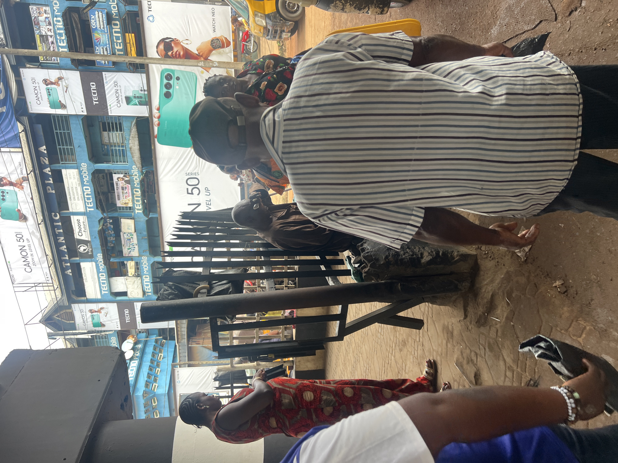 A street scene in an urban area, possibly Africa, shows a group of people gathered outside a building covered with Tecno Mobile advertising banners. The individuals are engaged in conversation or waiting, some seated on makeshift wooden structures. The environment is a bustling street with other businesses and passing traffic visible in the background. The weather appears to be sunny and the overall atmosphere is that of a busy commercial area.