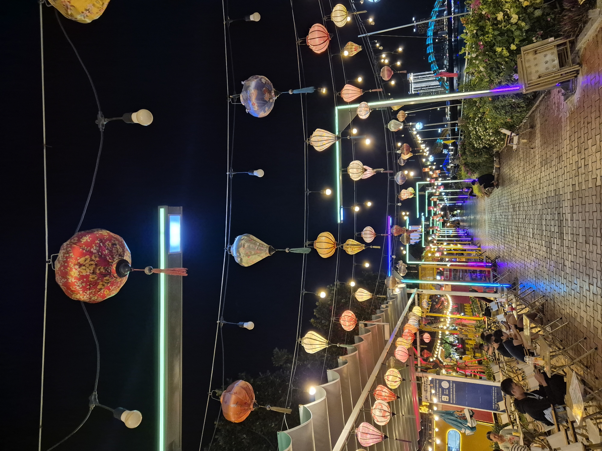 The image captures a vibrant nighttime scene along the Han River in Đà Nẵng, Vietnam. A pedestrian walkway, paved with brick, stretches into the distance, lined with an array of colorful hanging lanterns and string lights. The lanterns, predominantly red, orange, and yellow, create a festive atmosphere. To the left, a row of outdoor seating areas with tables and chairs suggests a dining or gathering space, with several individuals seated and engaged with their phones or conversing. Further down the walkway, more seating is visible, along with a vibrant display of neon lights in shades of blue, pink, and green. A lush green hedge borders the walkway on the right, and beyond that, the Han River is visible, reflecting the city lights. In the background, a modern cityscape is illuminated, featuring tall buildings and a distinctive bridge adorned with blue lights, likely the Dragon Bridge, a prominent landmark in Đà Nẵng. The overall impression is one of lively urban nightlife set against the backdrop of the river and illuminated architecture.