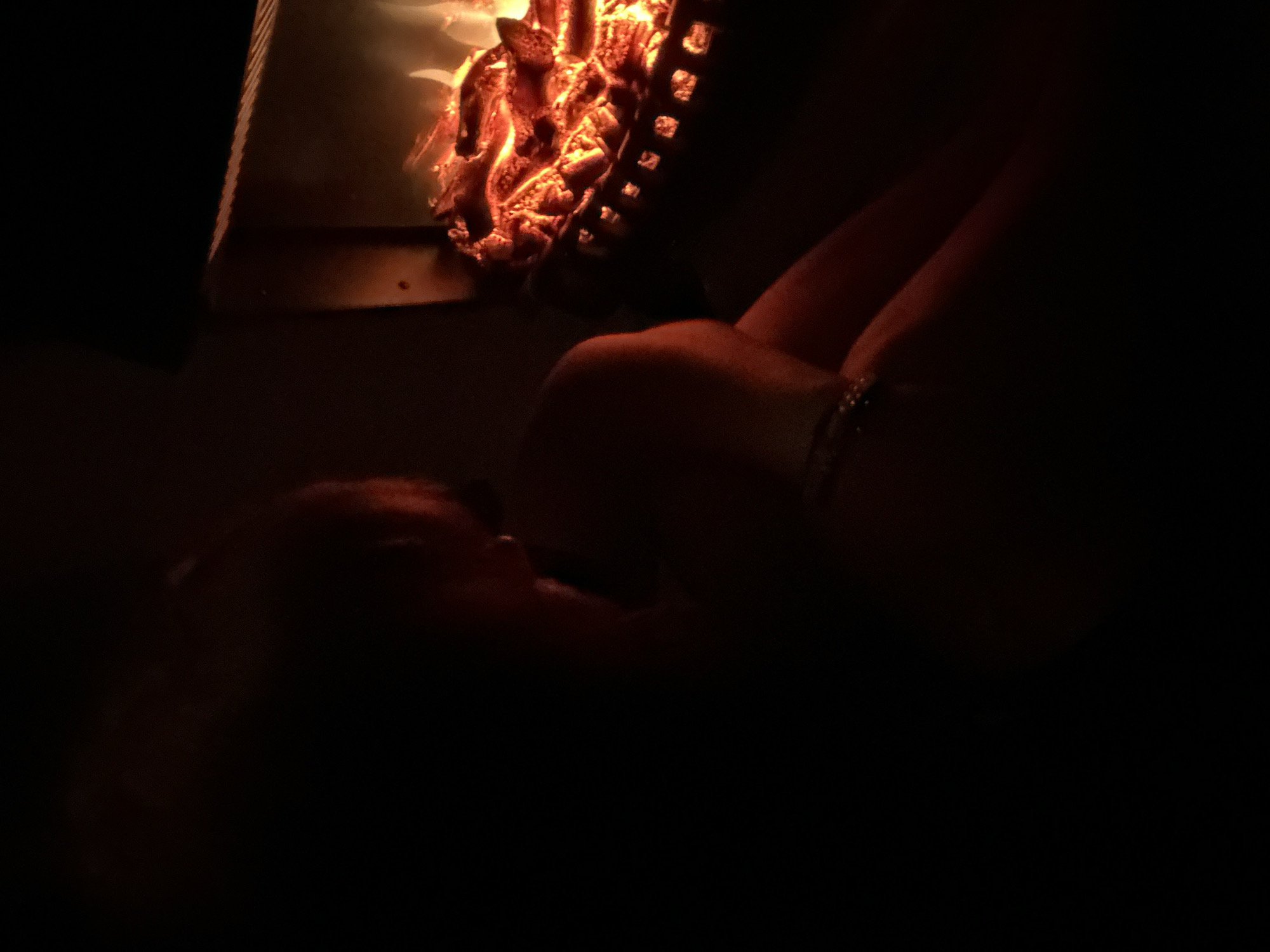 A person is partially visible in a dark room, positioned to the left of a fireplace with a glowing fire. The person is leaning in, with their head obscured in shadow, and appears to be wearing a bracelet on their left wrist. The fireplace is in the background, with the flames illuminating the logs and the metal grate. The overall scene is dimly lit, with the fire being the primary source of light. The setting is indoors, likely a home.